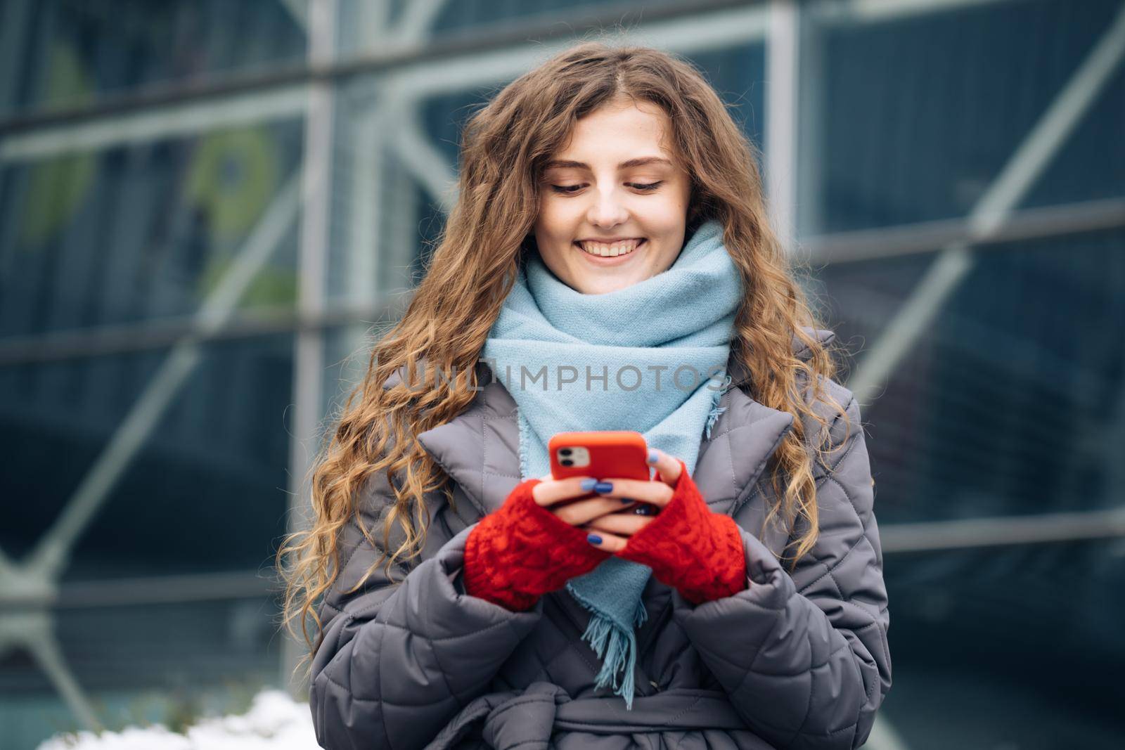Cheerful Curly-haired Young female texting on smartphone standing on street in winter city on New Year. Female tapping on cellphone outdoors. Online shopping by uflypro