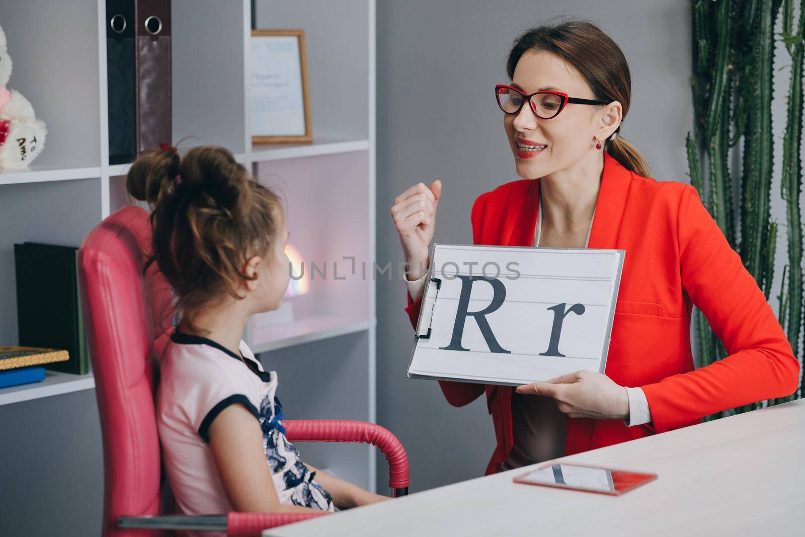 Speech therapist teaches the girls to say the letter R. Speech therapist holding letter R and girl back view.