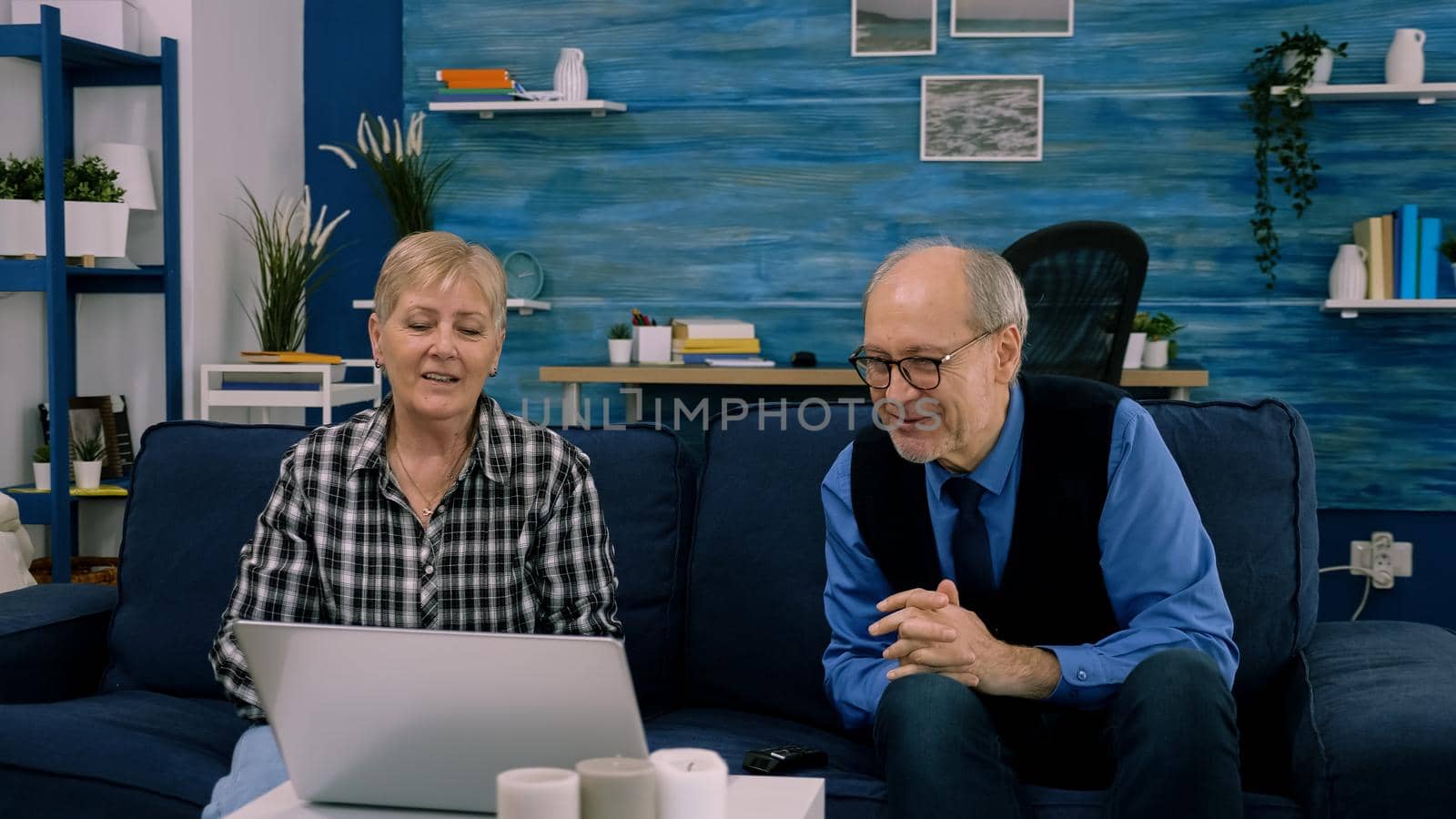 Elderly age couple sitting on sofa waving at camera during videocall by DCStudio