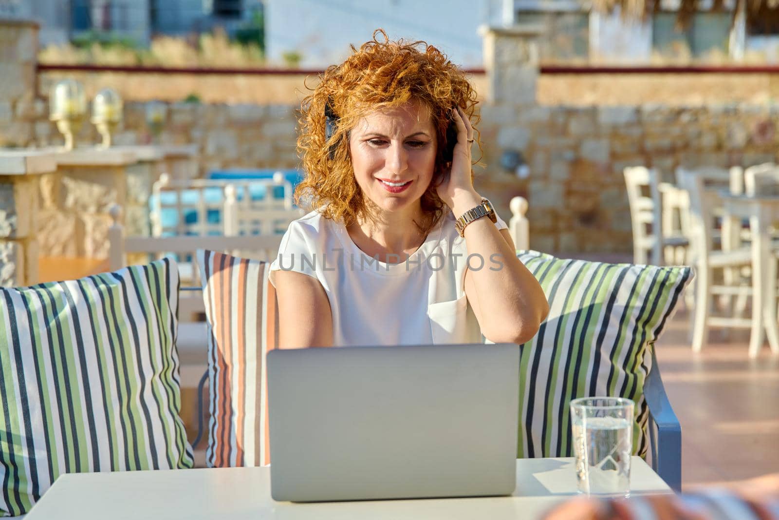 Positive smiling woman working with laptop outdoors. Middle aged female with headphones sitting at table in cafe, looking at monitor. Remote work, freelance, online business, video conference, chat