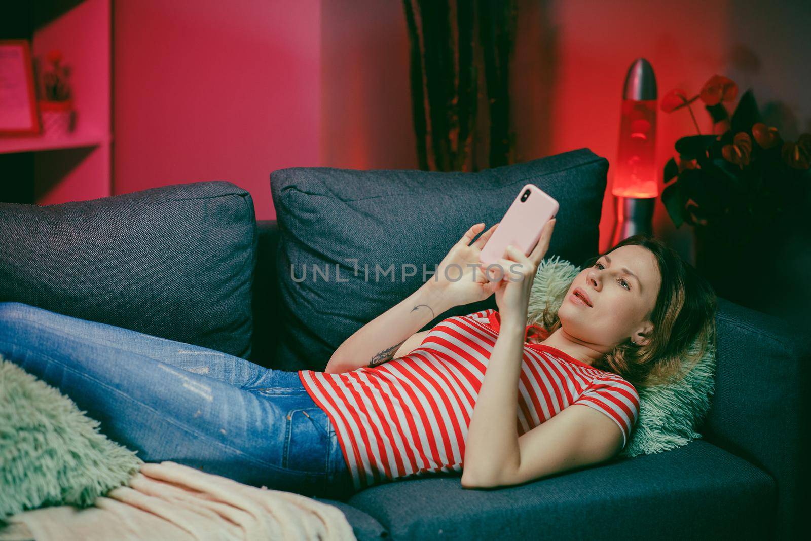 Relaxed girl holding smart phone using mobile apps watching funny video laughing lying on couch, smiling lazy young woman having fun chatting in social media resting on sofa at home by uflypro