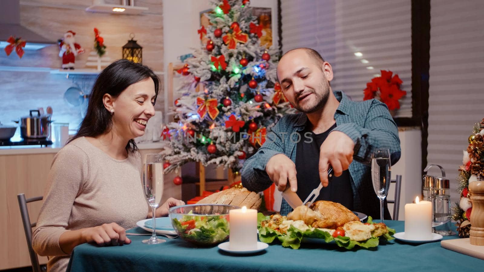 Man cutting chicken for woman at festive dinner on christmas eve festivity. Cheerful couple enjoying traditional meal and drinking champagne from glasses on table. Seasonal celebration