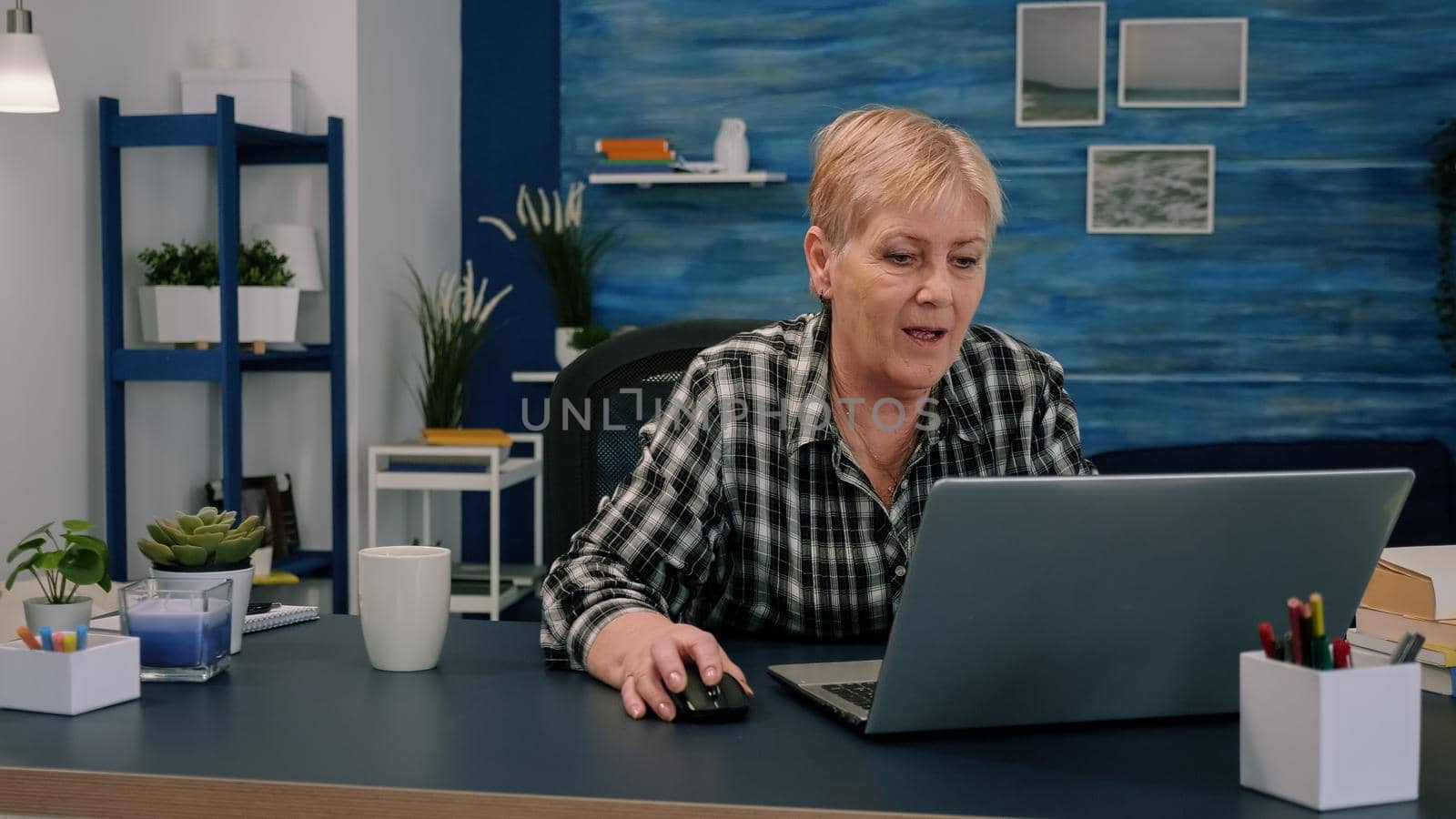 Old middle aged businesswoman working at laptop typing financial data sitting at desk using modern technology while retired husband sitting on couch. Serious lady analyzing and managing domestic bills