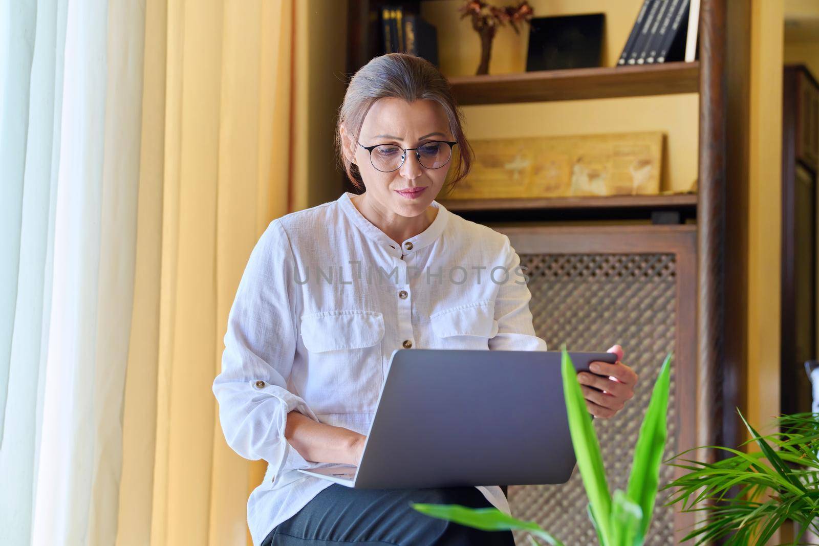 Middle-aged woman psychologist in an office with laptop. Portrait of professional female counselor, psychiatrist, therapist, teacher sitting on chair near window. Technology, medicine, people