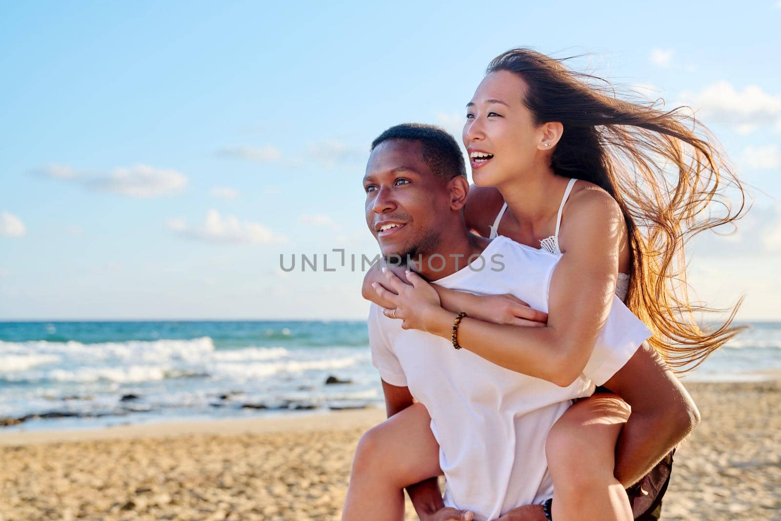 Young beautiful having fun couple on sea background. Multiracial couple, asian woman on back of african american man, vacation together, relationship dating happiness, happy lovers moments, copy space