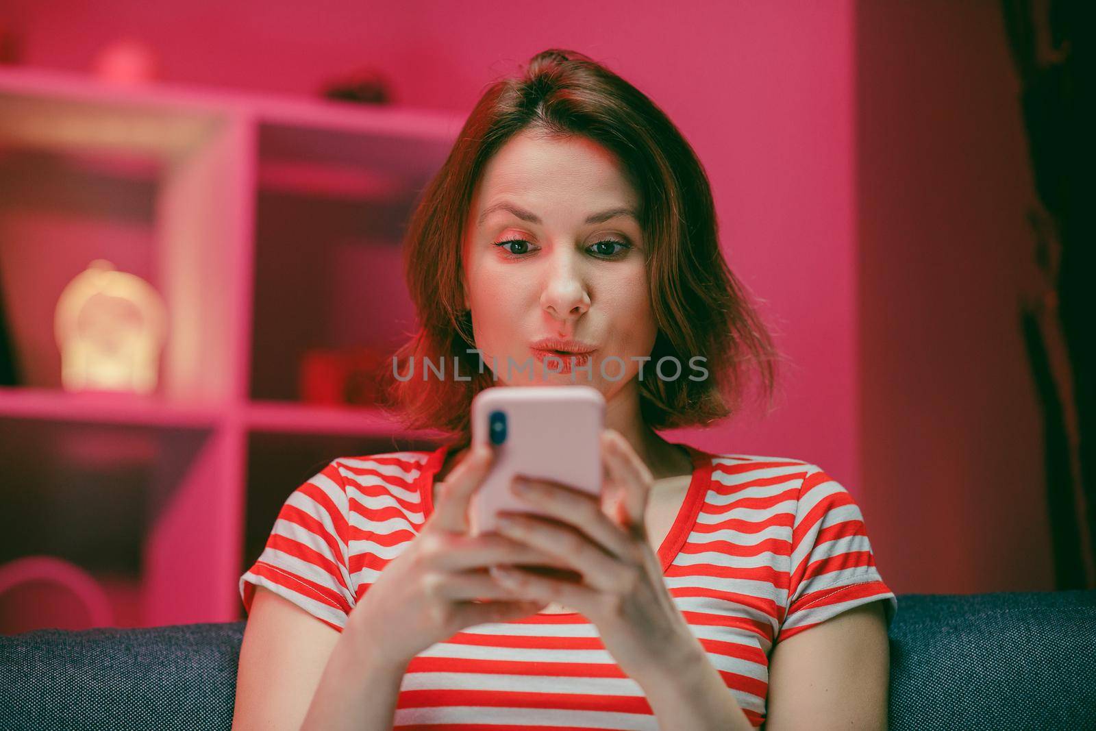 Happy Young Woman Holding Smart Phone Looking at Cellphone Screen Laughing Enjoying Using Mobile Apps for Shopping. Having Fun Playing Games Chatting in Social Media Sit on Couch at Home. by uflypro