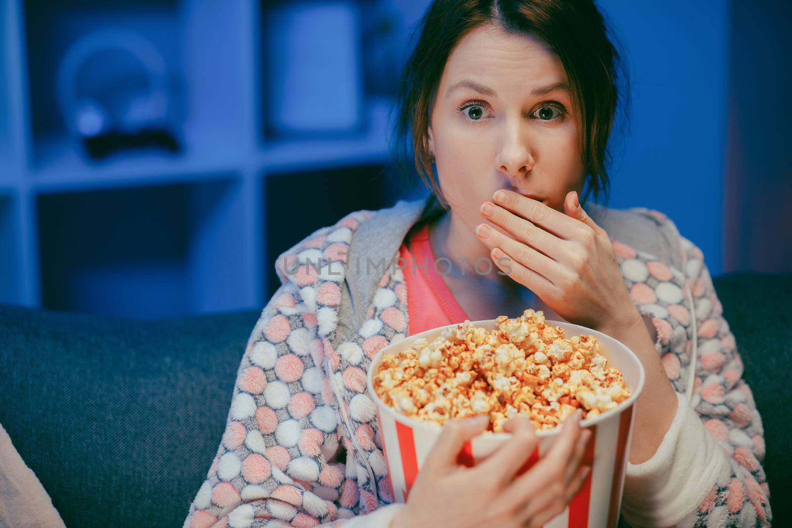 Woman with popcorn sitting on the sofa watching something scary while eating popcorn and being afraid. Inside