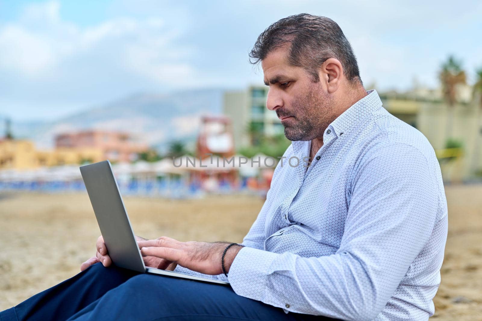 Serious confident mature man with laptop outdoors. Business male working with laptop sitting on sand at beach. Remote business, technology, freelance, lifestyle, leisure, middle age concept