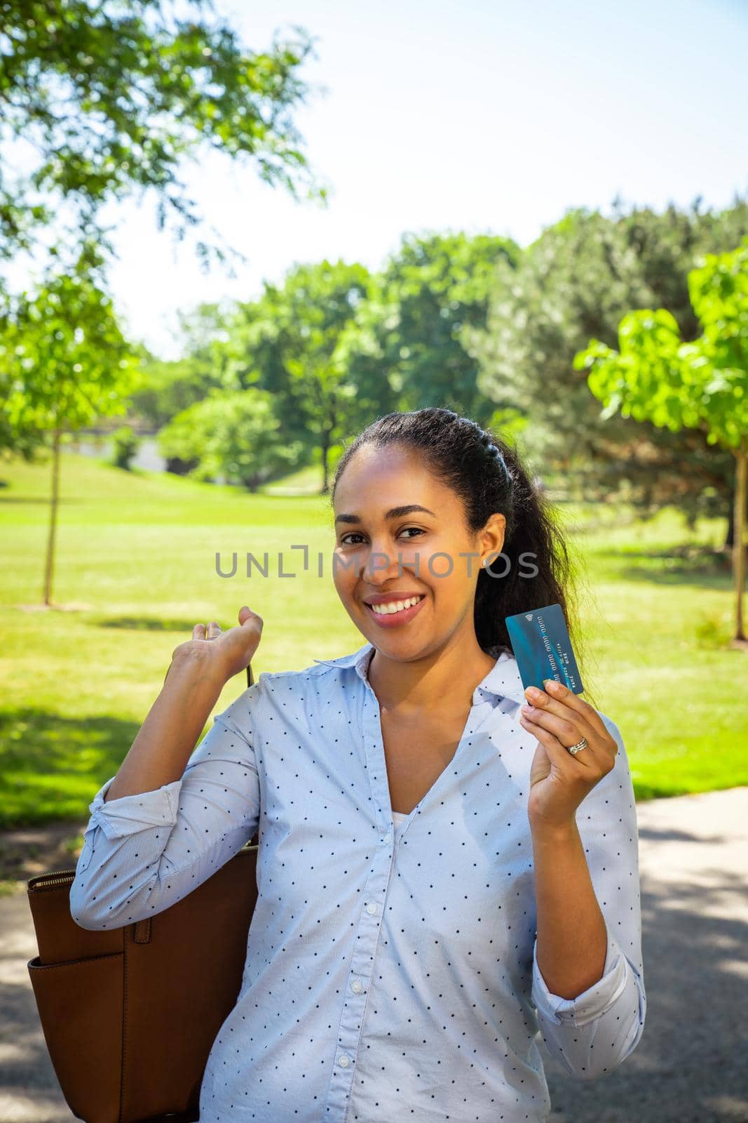 A smiling mixed race African American woman in a blue collared shirt holds up a blue plastic credit card with micro chip with her brown purse slung over a shoulder outside in a park. by lapse_life