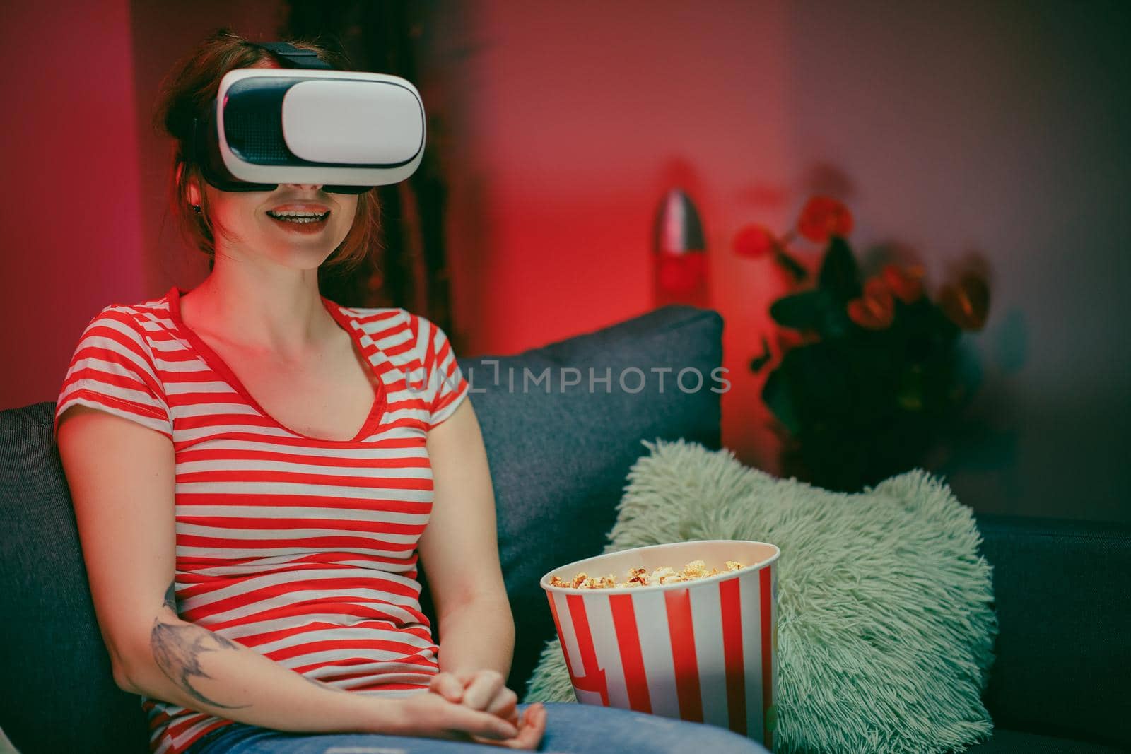 Portrait of the young woman sitting on the couch and having the VR headset, watching something while eating popcorn and smiling. Indoors