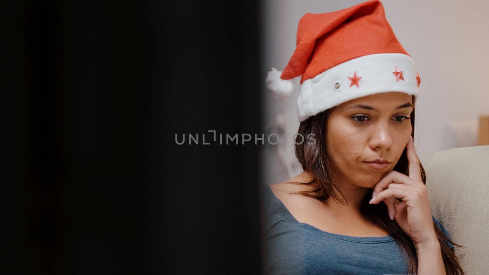 Close up of unhappy person working on laptop at television. Festive woman wearing santa hat using modern device on christmas eve. Adult laying on couch with gadget on holiday.