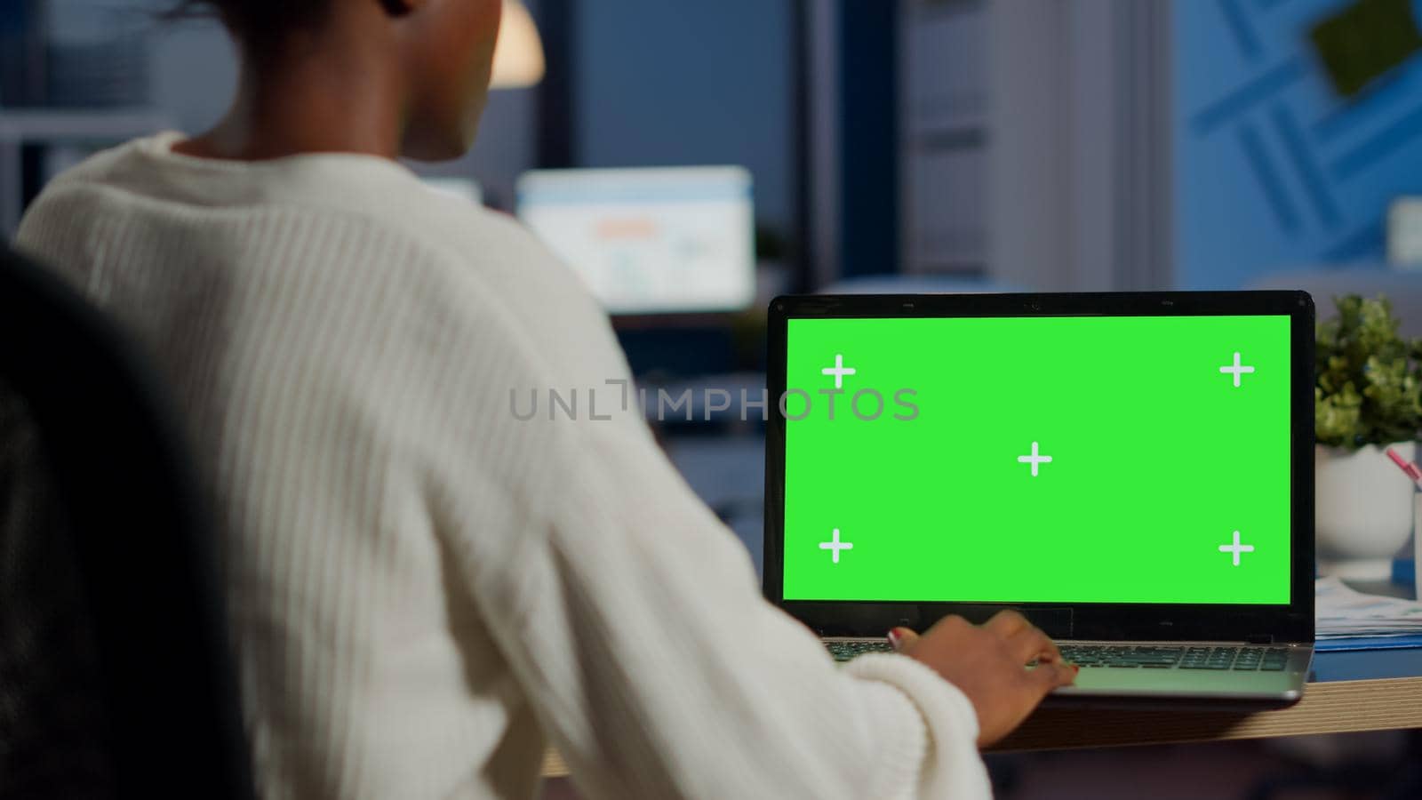African woman manager looking at mock up monitor of laptop, green screen mockup, chroma key desktop sitting at desk in business office late at night. Freelancer watching at isolated desktop overwork