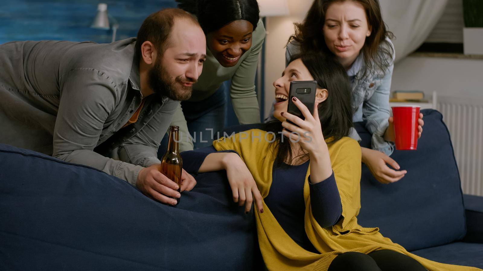 Group of mixed race people spending time together while looking at comedy video on phone. Group of mixed race people sitting on sofa, drinking beer, chilling in living room late at night