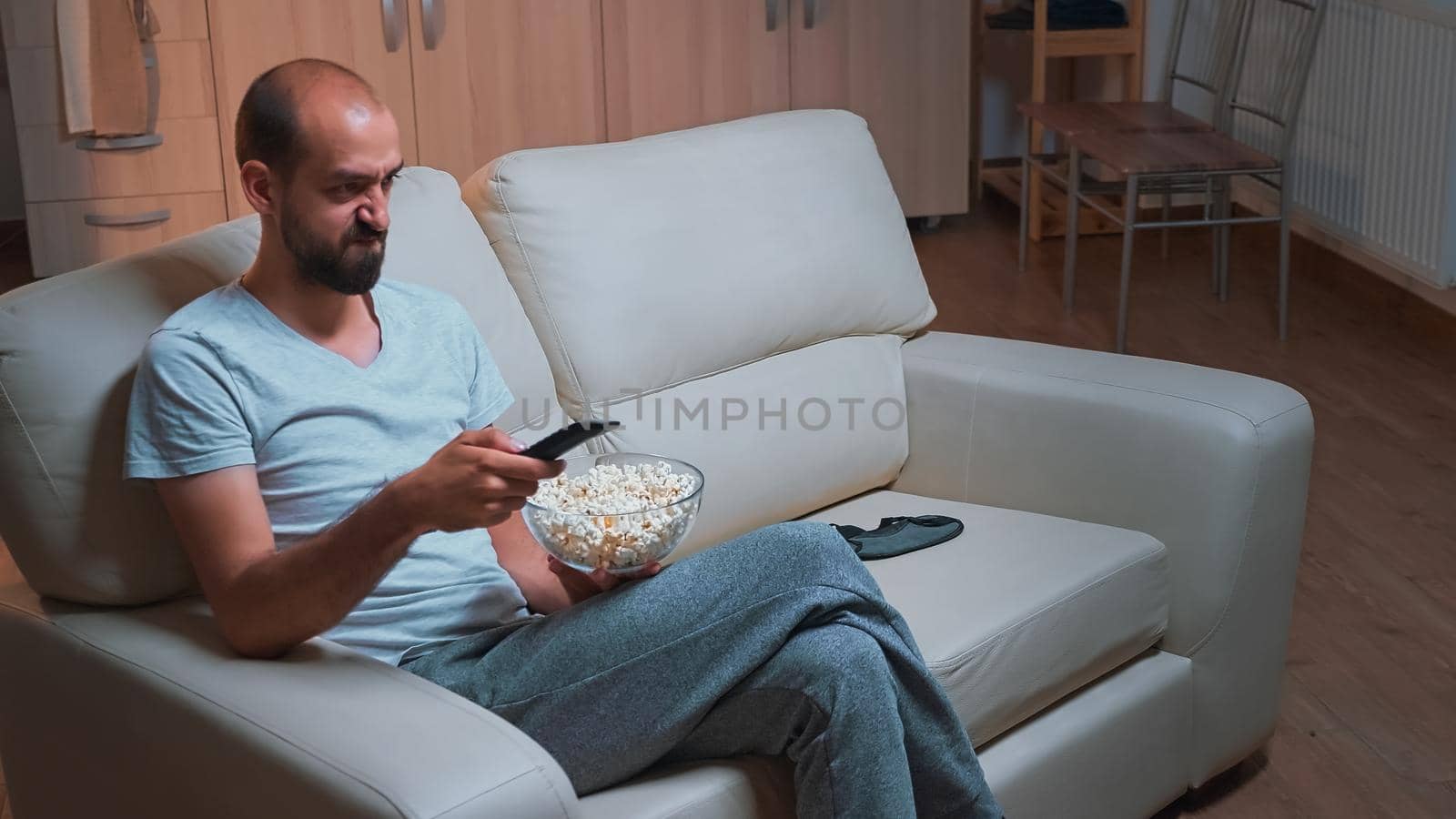 Concentrated man sitting in front of television using control remote while watching movie film. Caucasian male in pajamas looking at entertainment shows late at night in kitchen