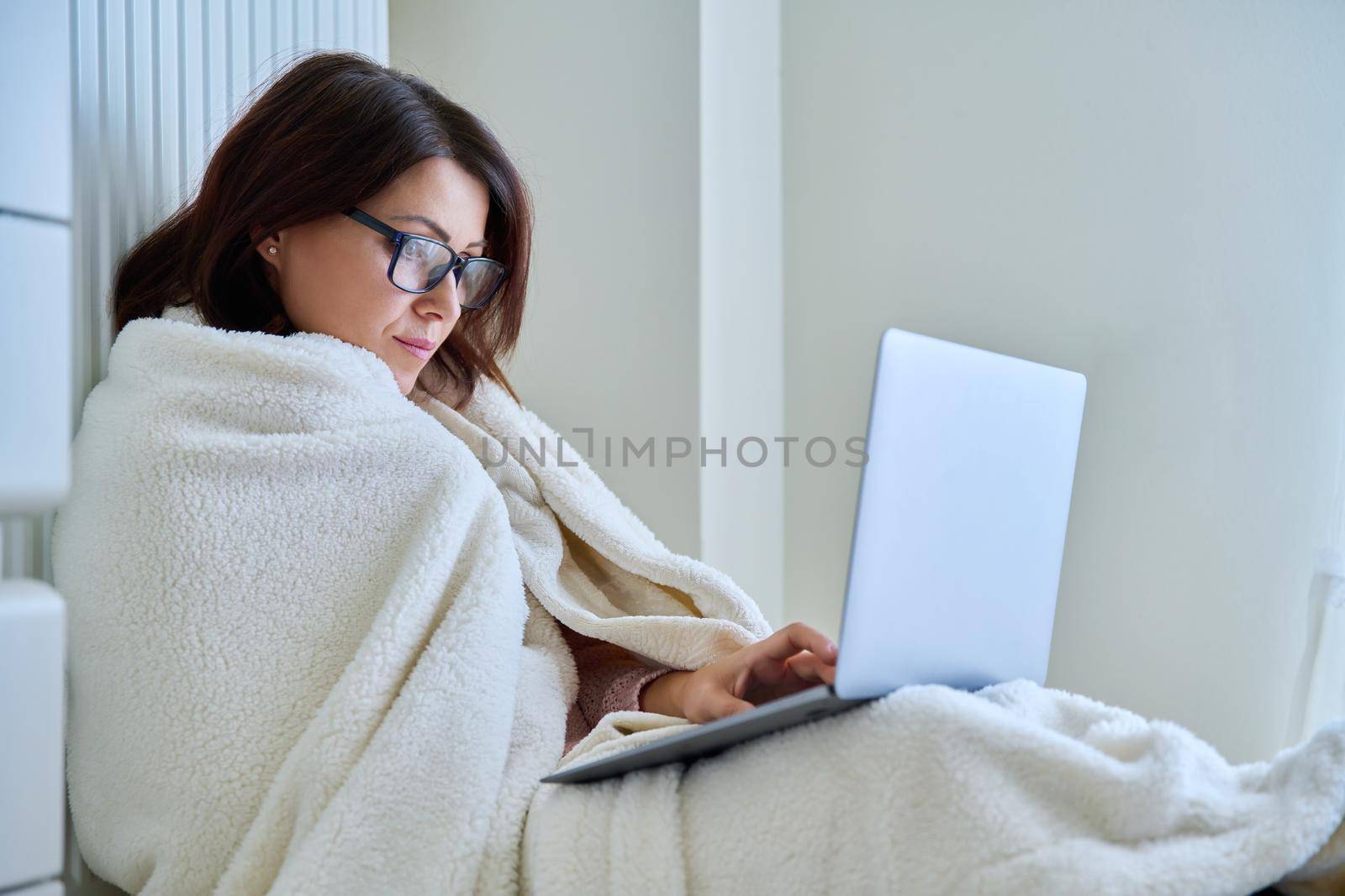 A frozen mature woman working at home with a laptop, warming herself with a blanket and heating radiator. Autumn winter cold season