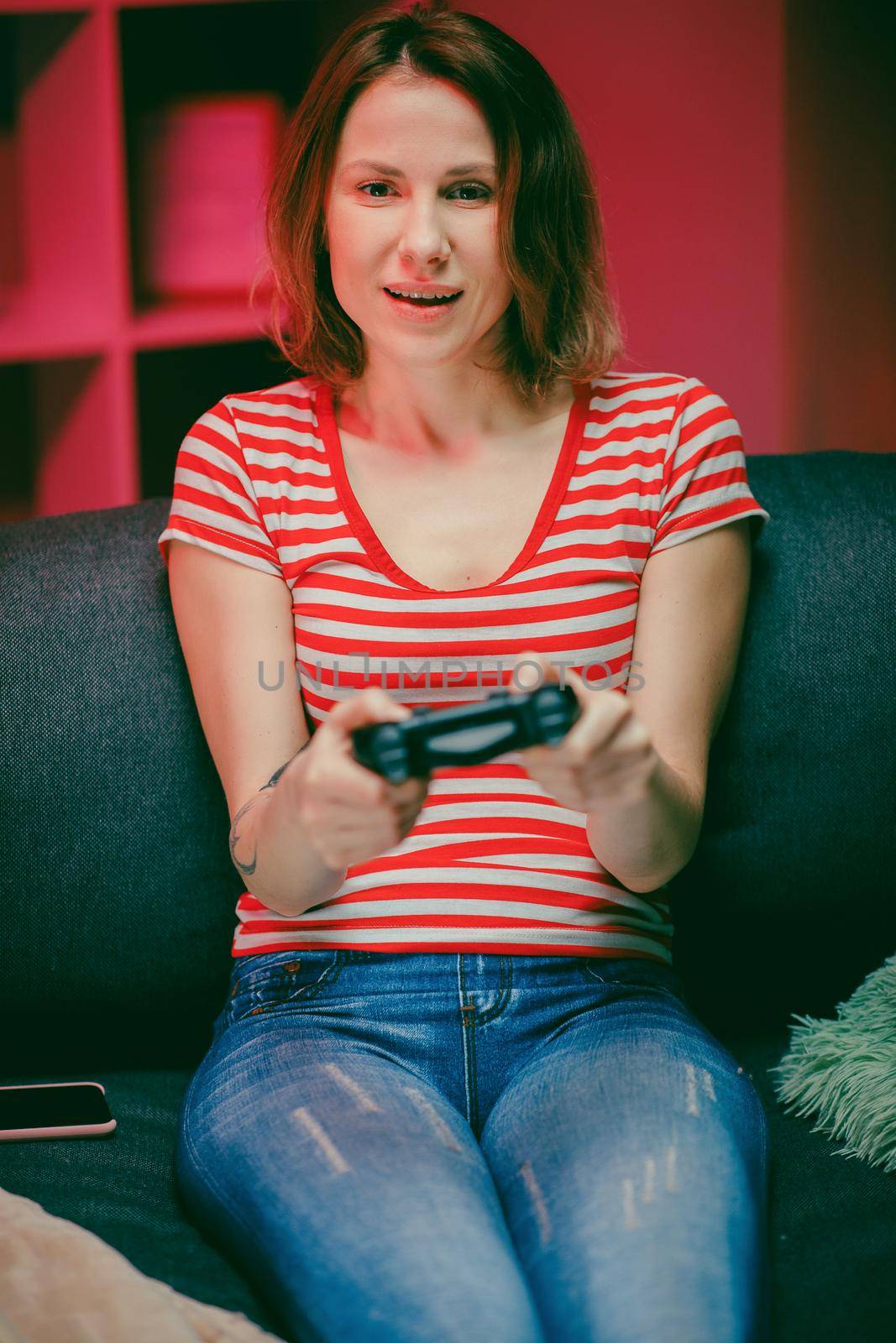 Girl playing a video game at living room at night. Gamer woman sitting on a sofa, playing in video games on a console and using a wireless controller.