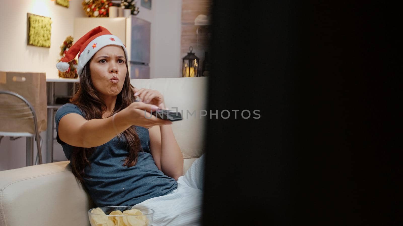 Frustrated person switching channels on TV using remote control, feeling angry on christmas eve holiday. Woman with santa hat and decorated room looking at movie on television with chips