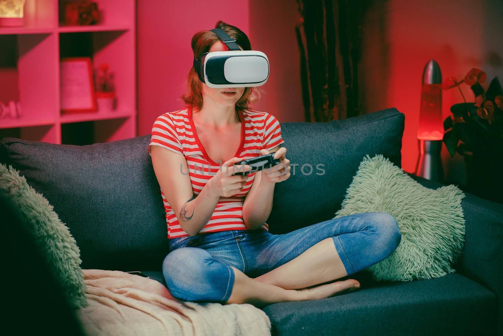 Young woman testing virtual reality glasses while sitting on sofa in home interior. Caucasian female with vr headset on face playing game with smile on couch in modern apartment