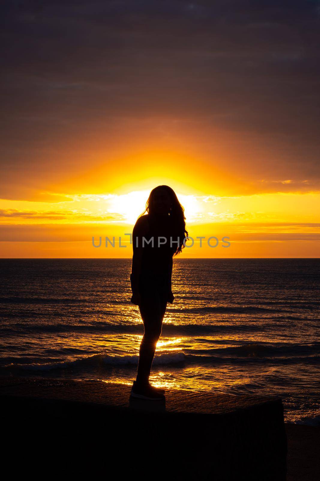 A gorgeous silhouette of a woman with long hair standing on the beach with the glow from the sun illuminating her outline as orange and yellow colors reflect off the water and cover the sky.