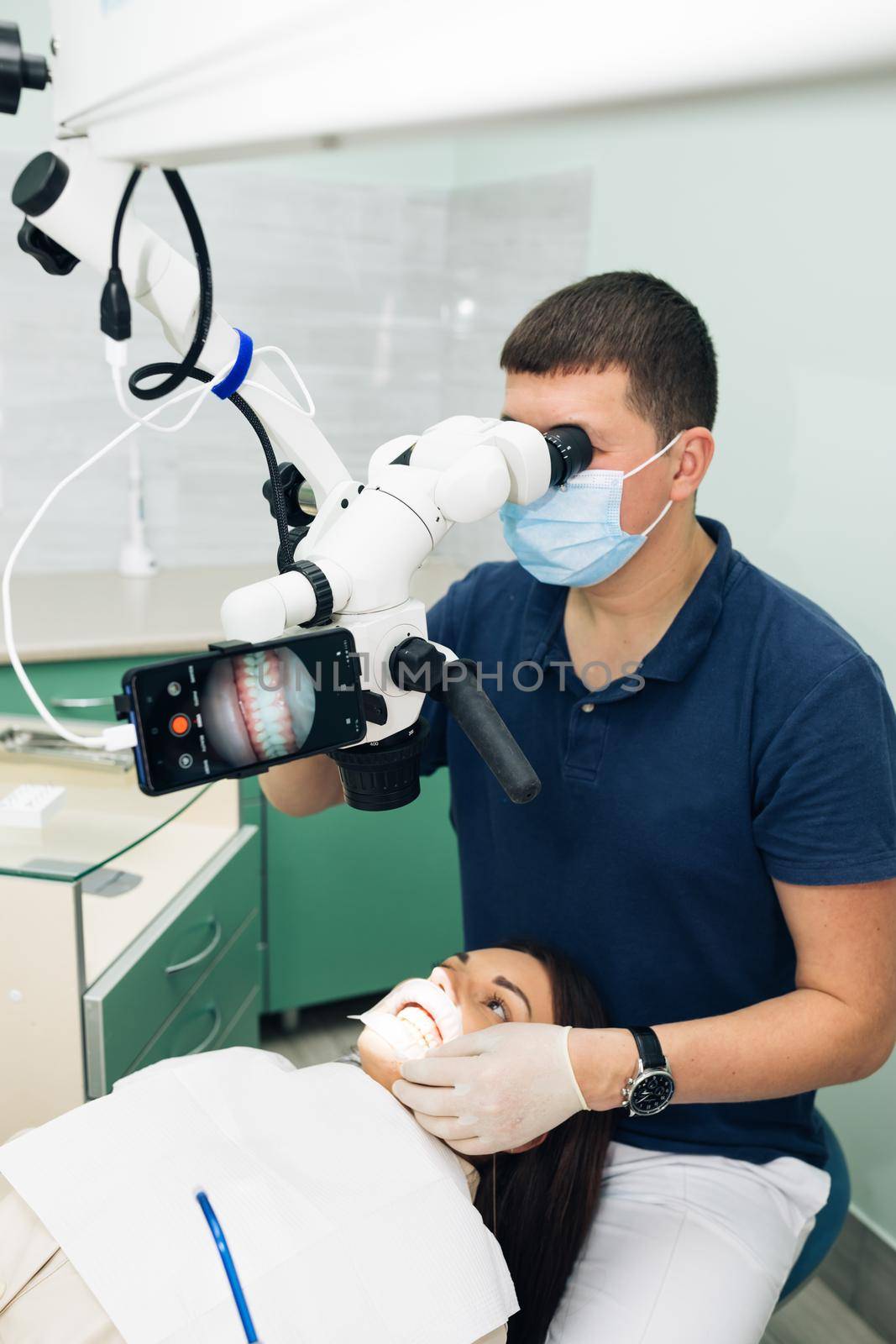 Dentist in latex gloves examining patient teeth in clinic. Patient lying with his open mouth in dentists office. Stomatologist performing examination using a microscope by uflypro
