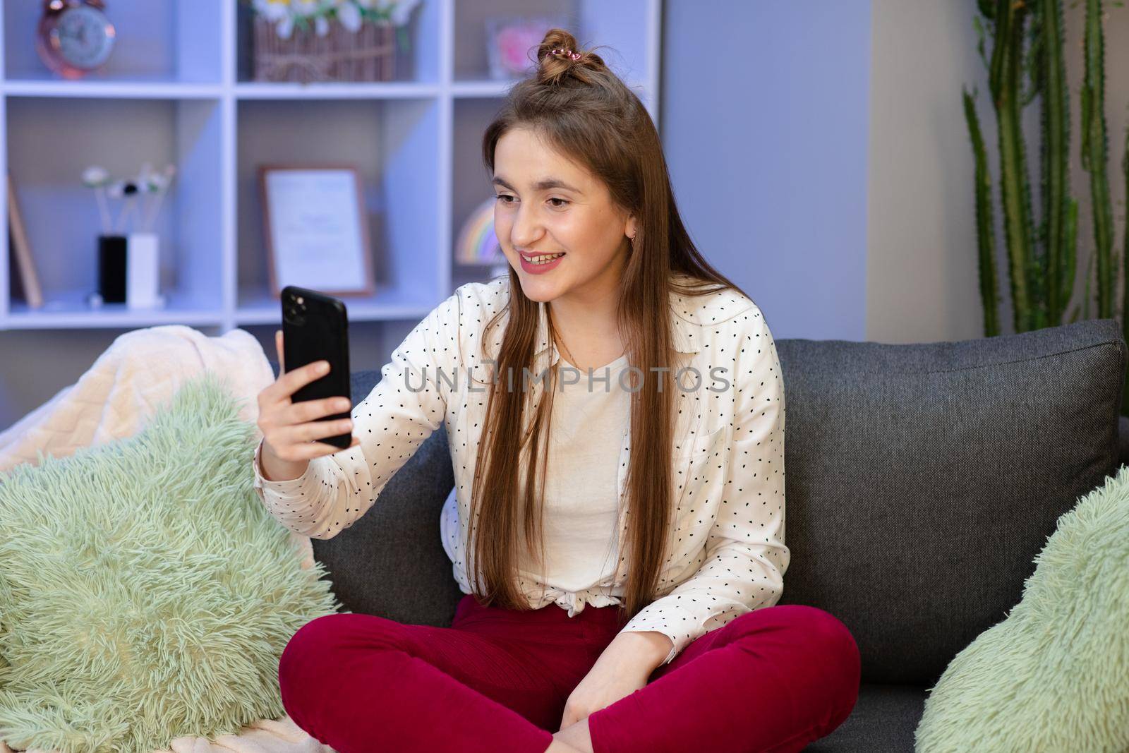 Happy young woman blogger influencer holding modern smart phone wave hand hello. Smiling vlogger girl looking at mobile make video call, shooting vlog taking selfie isolated on home background by uflypro