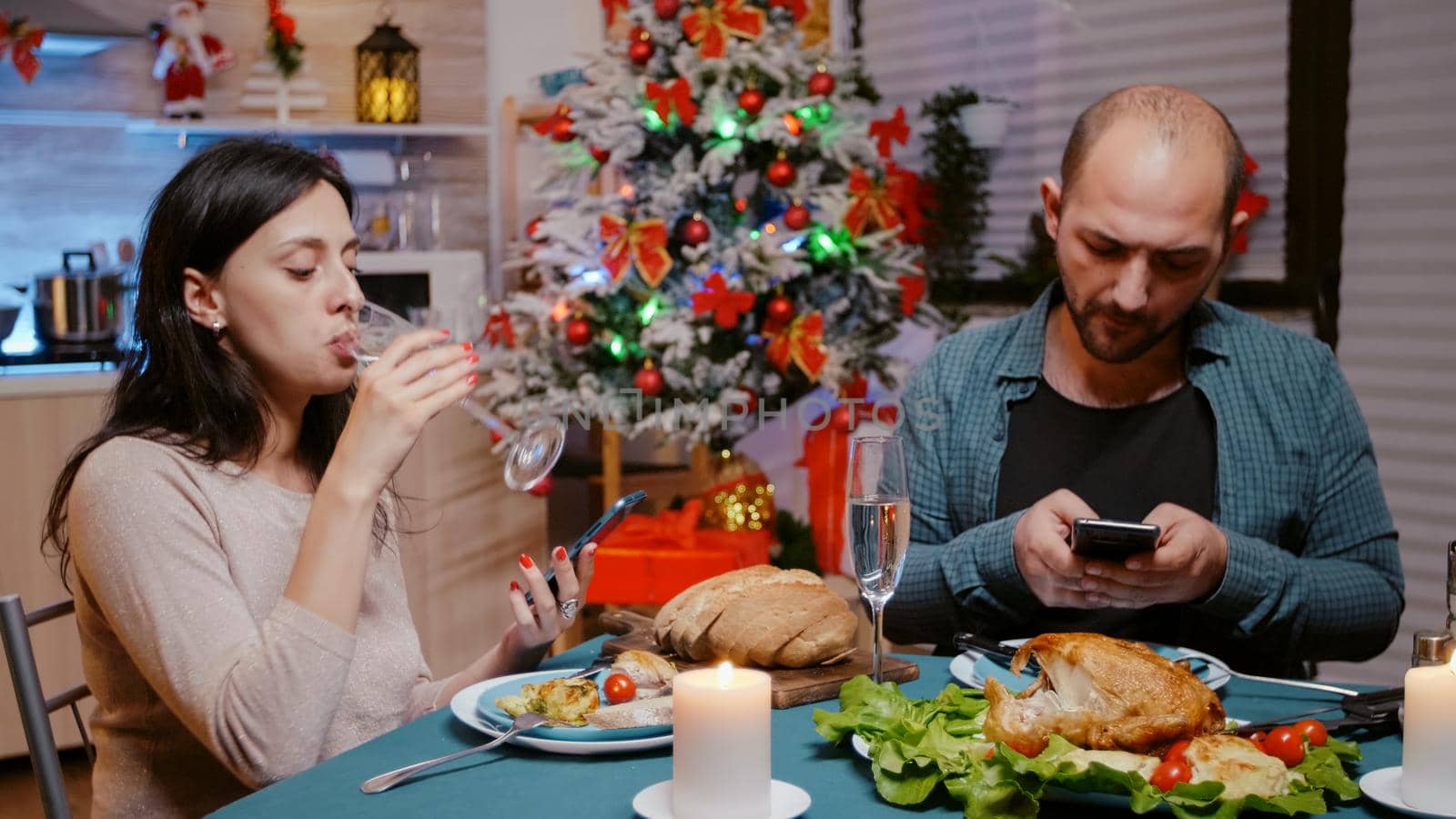 Couple holding smartphones and drinking champagne from glasses while celebrating christmas with festive dinner at home. Man and woman looking at devices after eating chicken meal.