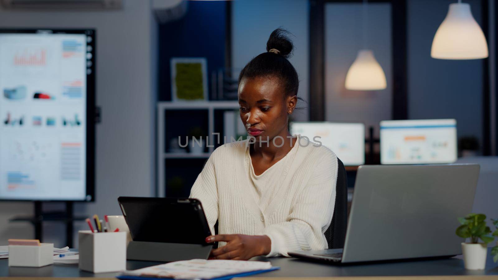 African manager woman using laptop and tablet in same time working overtime in business start-up office. Busy multitasking Busy employee analysing financial statistics overworking writing, searching.