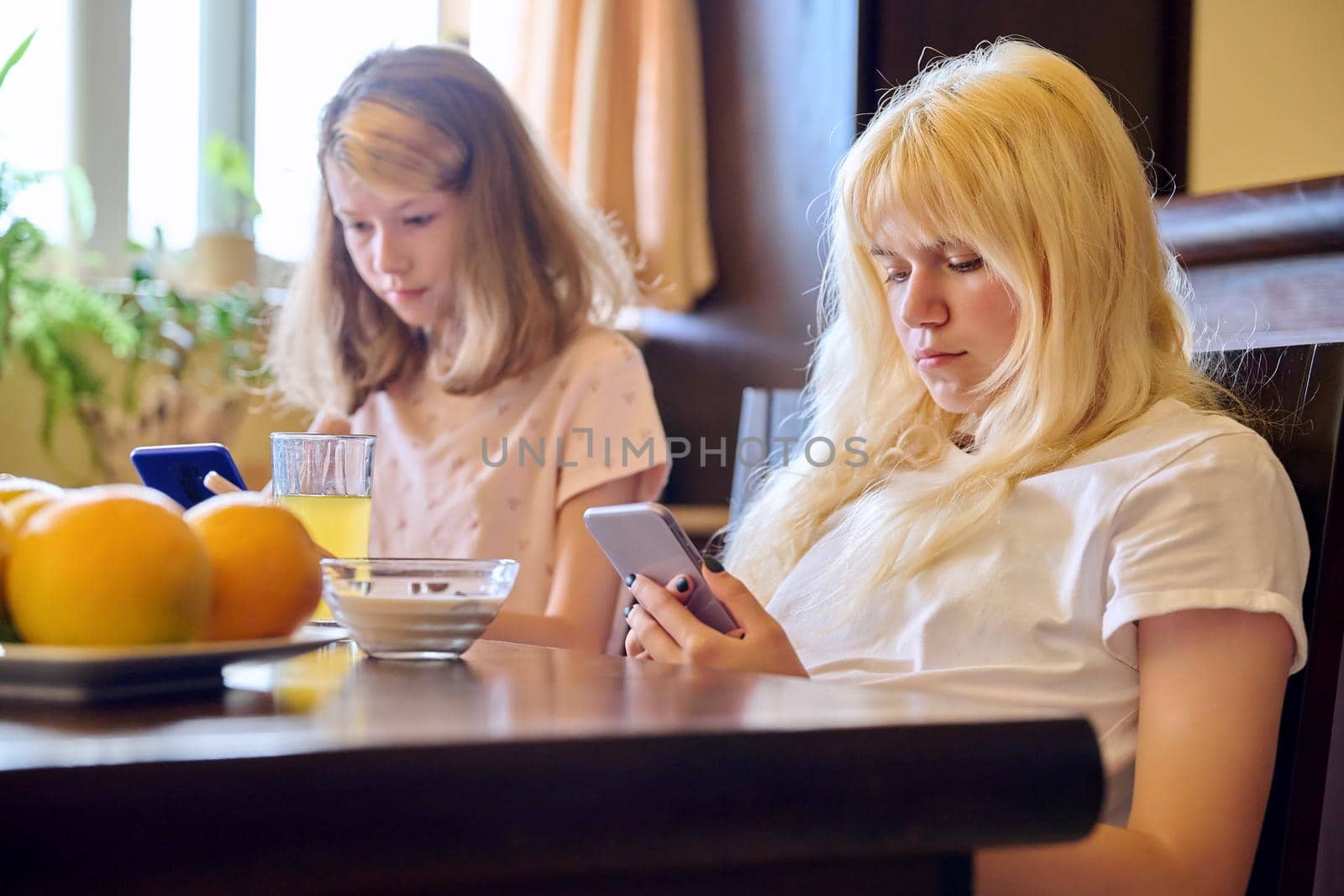 Children two girls sisters eating at home, looking at smartphones. Teenagers having breakfast in kitchen, using phones for leisure entertainment education. Technology in life lifestyle, adolescence