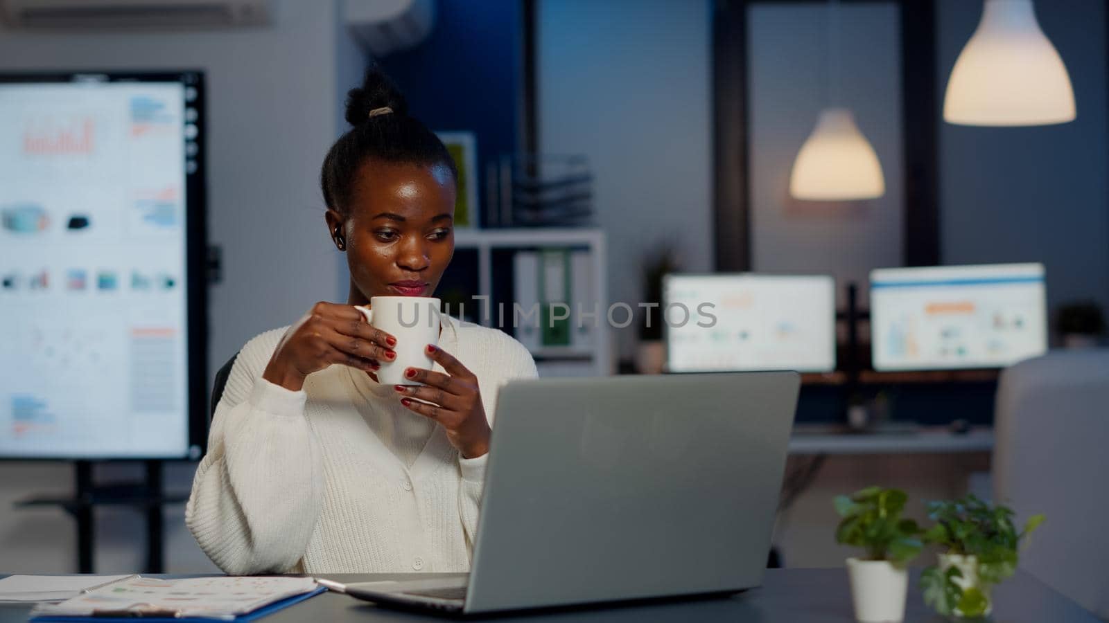 Black business woman using wireless headphone drinking coffee duing video conference working overtime from start-up office in front of laptop. Freelancer using talking on virtual meeting at midnight