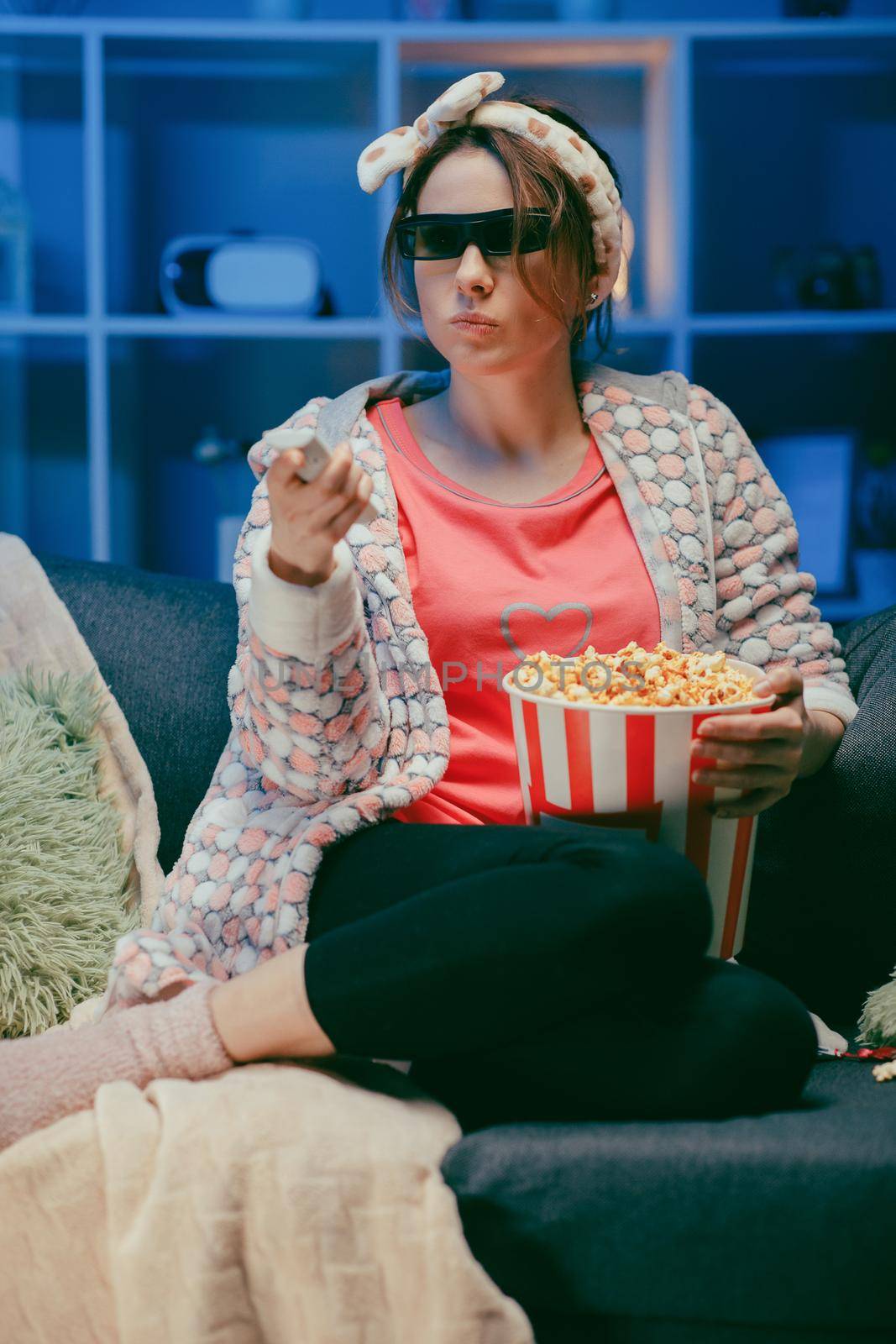 Woman on a soft couch eating popcorn, looking at the TV screen in 3D glasses. Woman eating popcorn in 3d glasses. by uflypro