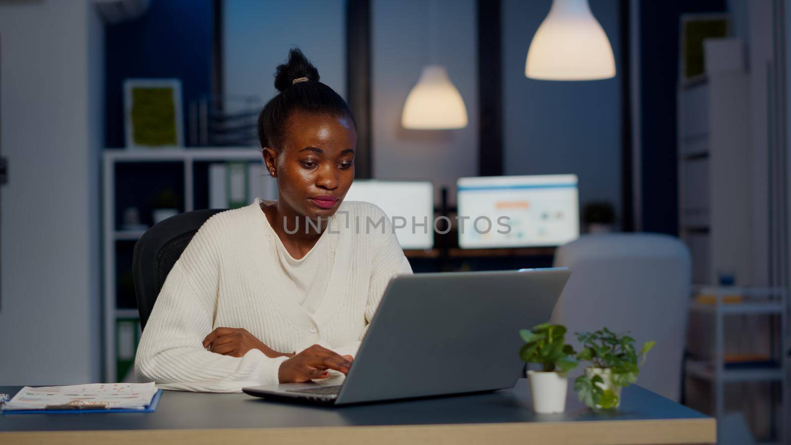 Stressed african manager woman working with financial graphs checking graphs, typing on laptop, reading raports late at night in start-up office doing overtime to respect deadline of project.