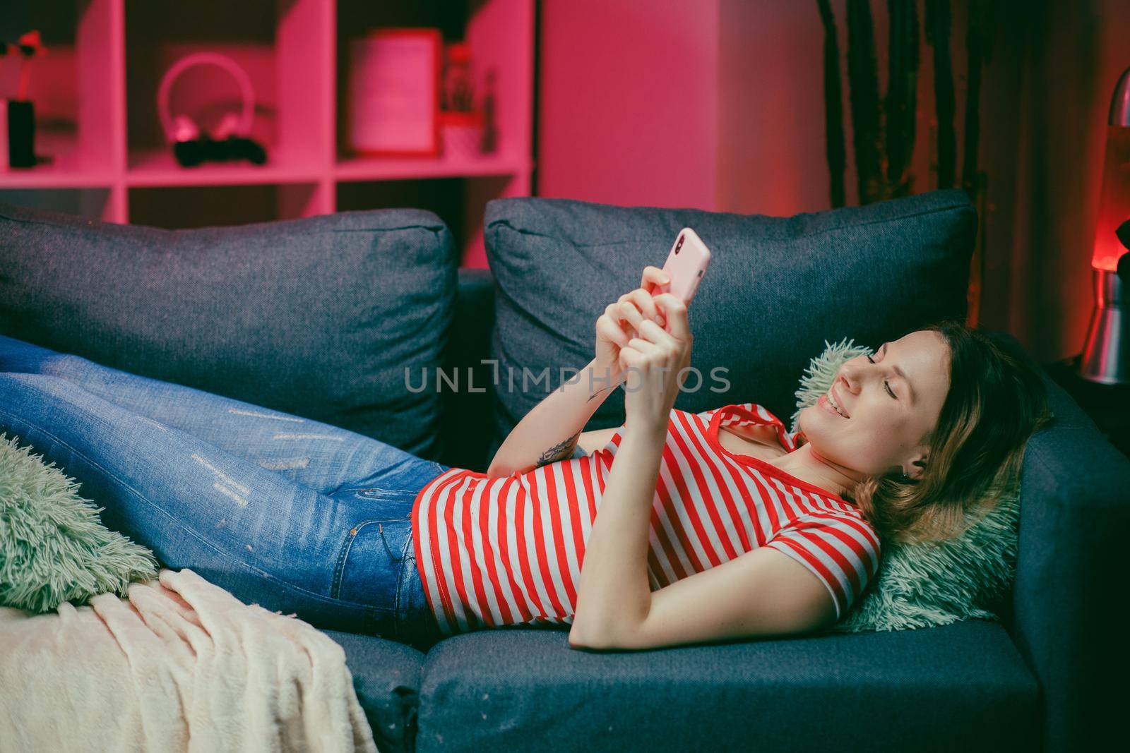 Relaxed girl holding smart phone using mobile apps watching funny video laughing lying on couch, smiling lazy young woman having fun chatting in social media resting on sofa at home