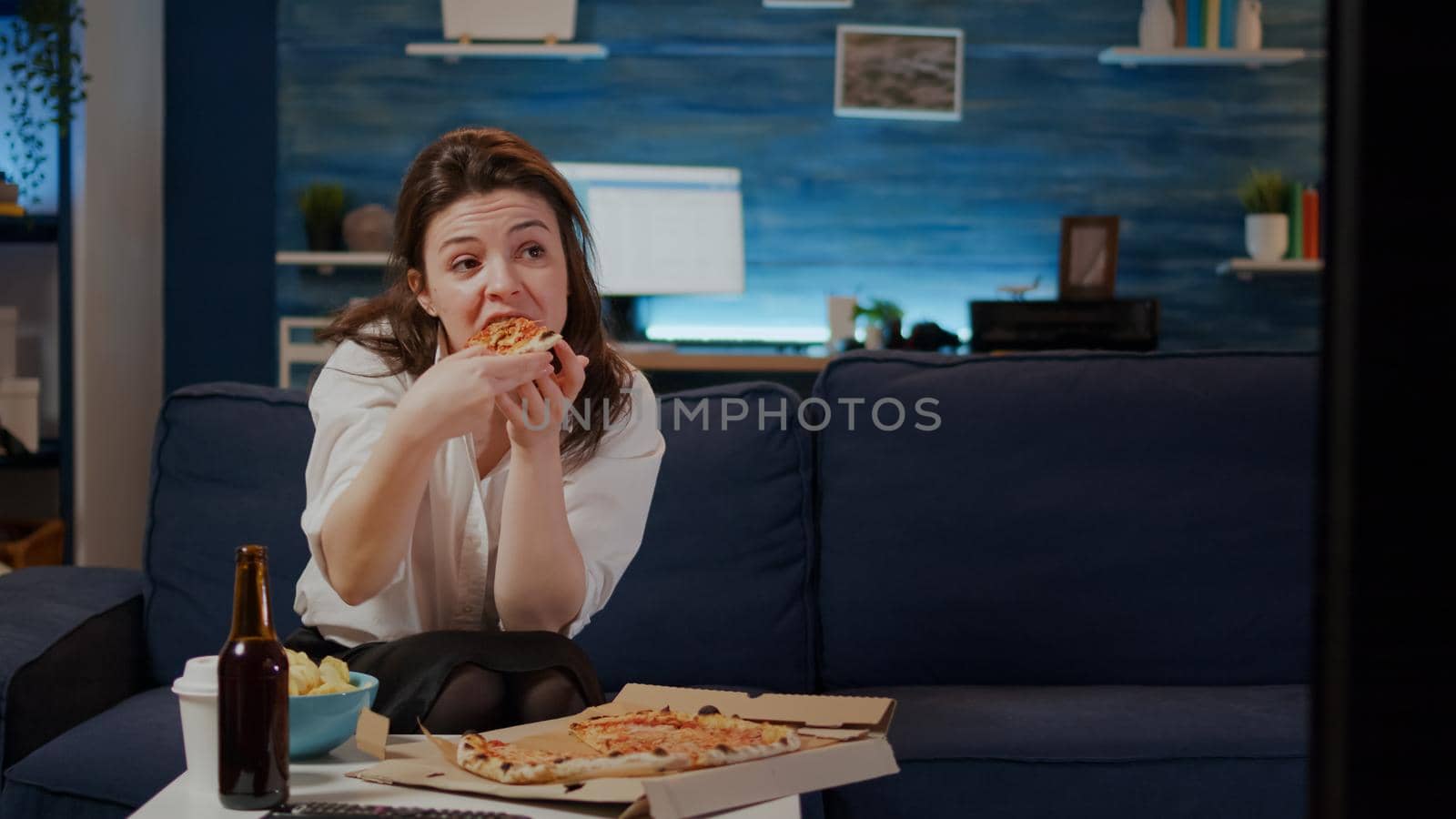 Young woman eating slice of pizza from box and drinking beer from bottle sitting in living room. Adult with fast food takeaway enjoying meal while looking at movie on television
