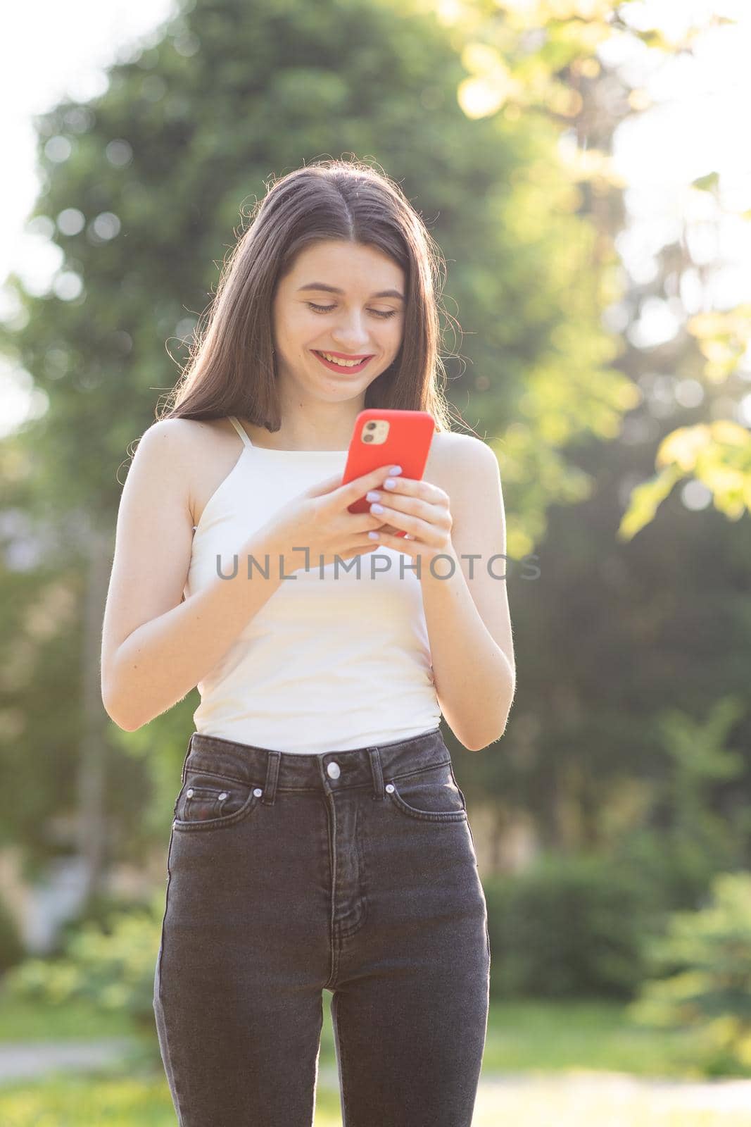 Girl is laughing and reading something on her Smartphone. Cheerful young woman communicating on smartphone while walking in park. by uflypro