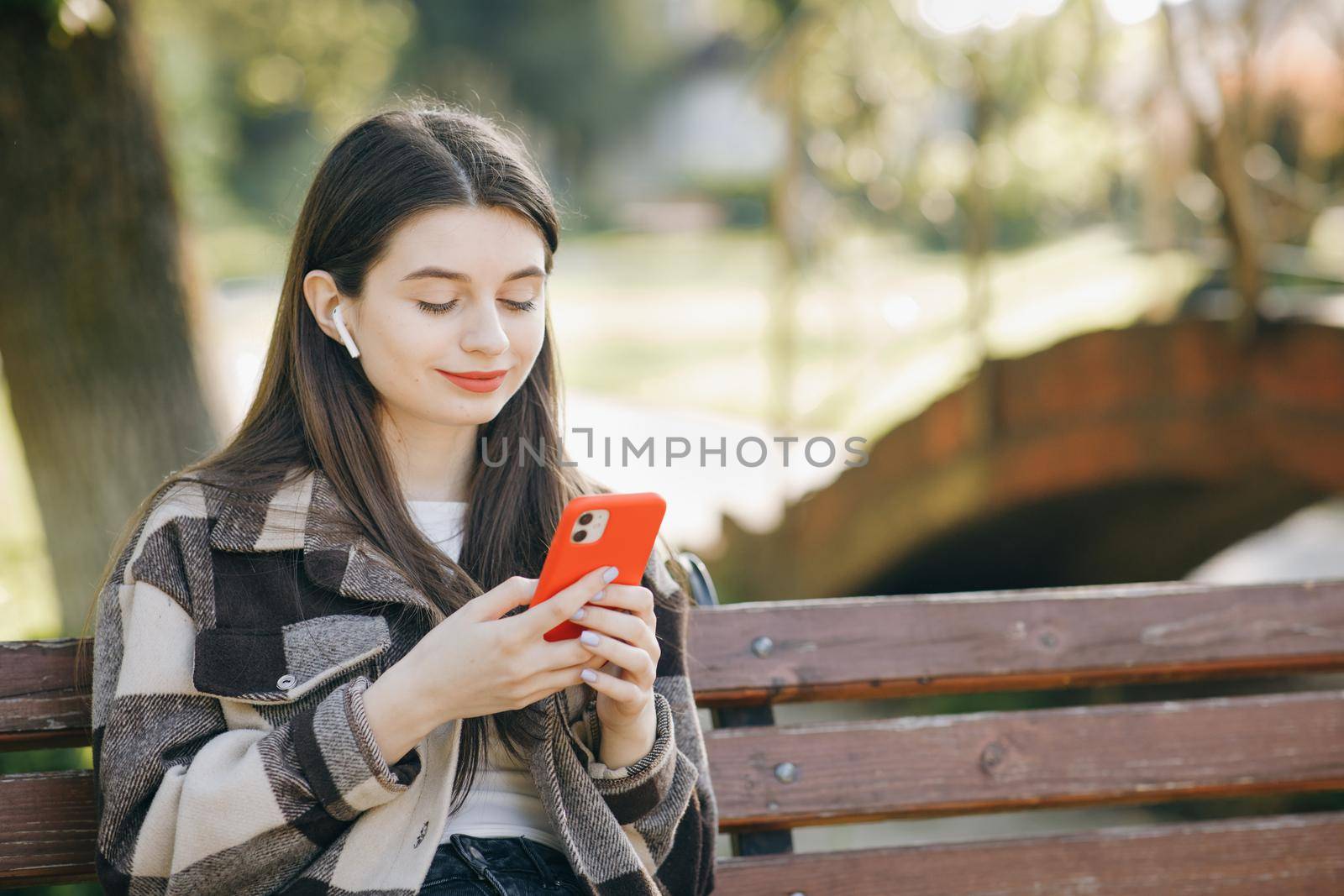 Attractive woman browsing on mobile phone in public park. City, urban background. Happy girl listening to music in the park using mobile phone and wearing headphones by uflypro