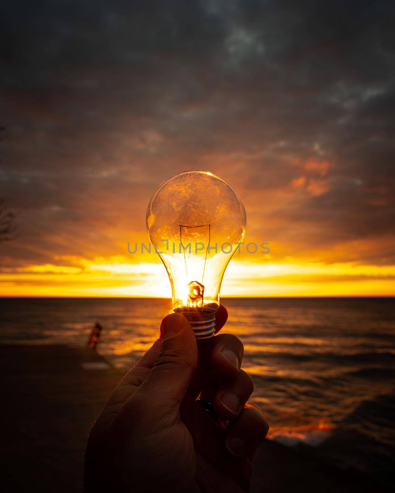 A man holds out a clear lightbulb in front of a colorful sunrise over Lake Michigan with rays of yellow, orange and pink filling the glass of the bulb making a beautiful background image.