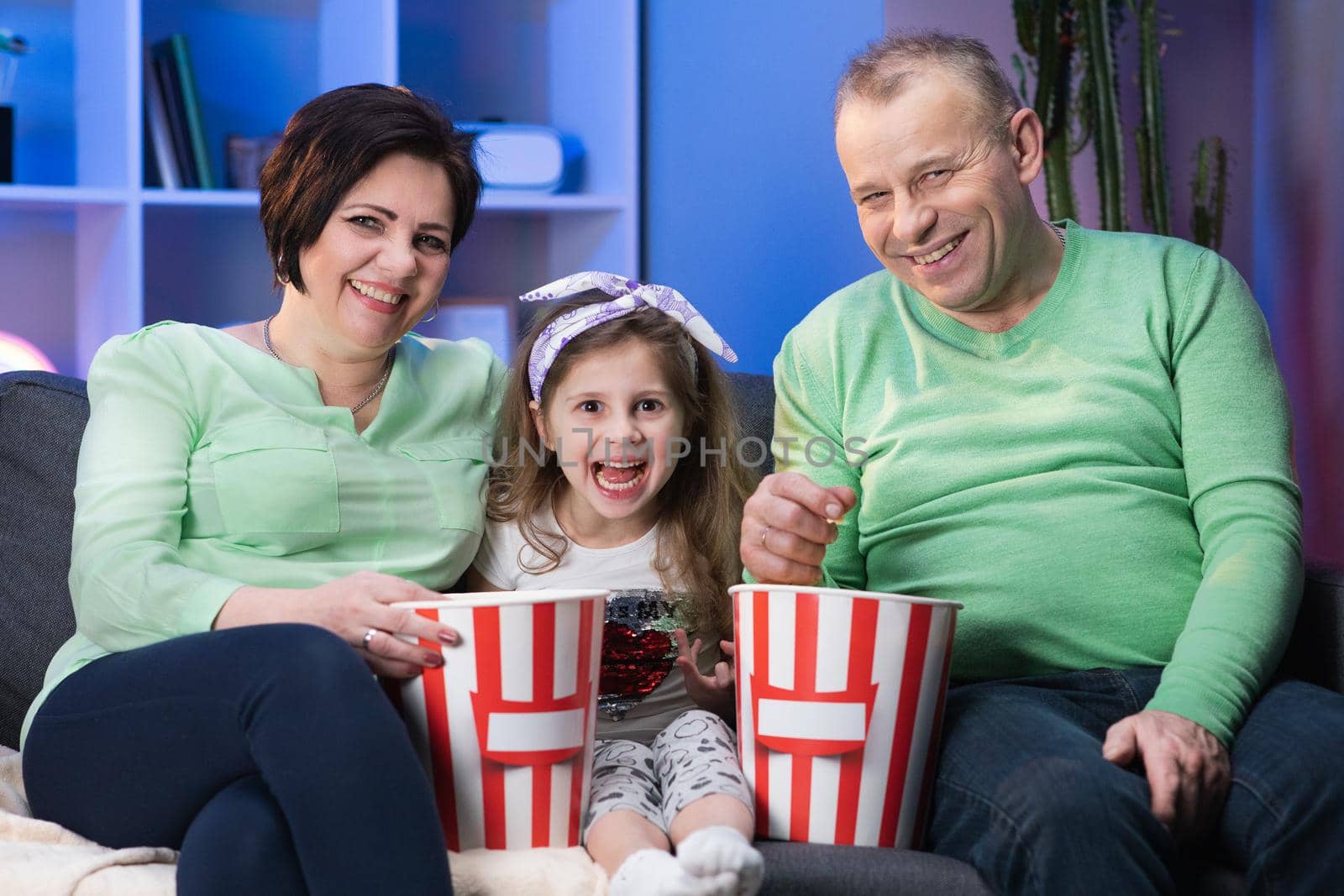 Happy family is sitting on sofa and relaxing by choosing and watching tv. Family, generation and people concept. Happy smiling grandmother, grandfather and little granddaughter