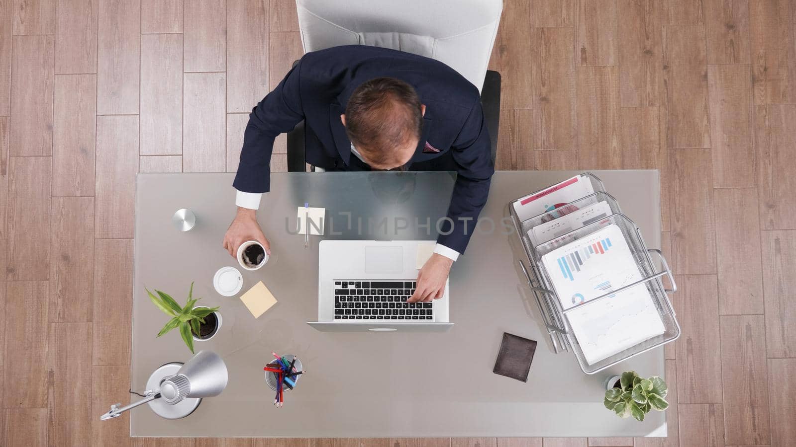 Top view of businessman holding cup of coffee while typing company statistics working at management strategy in startup office. Executive manager in suit analyzing investments documents on computer