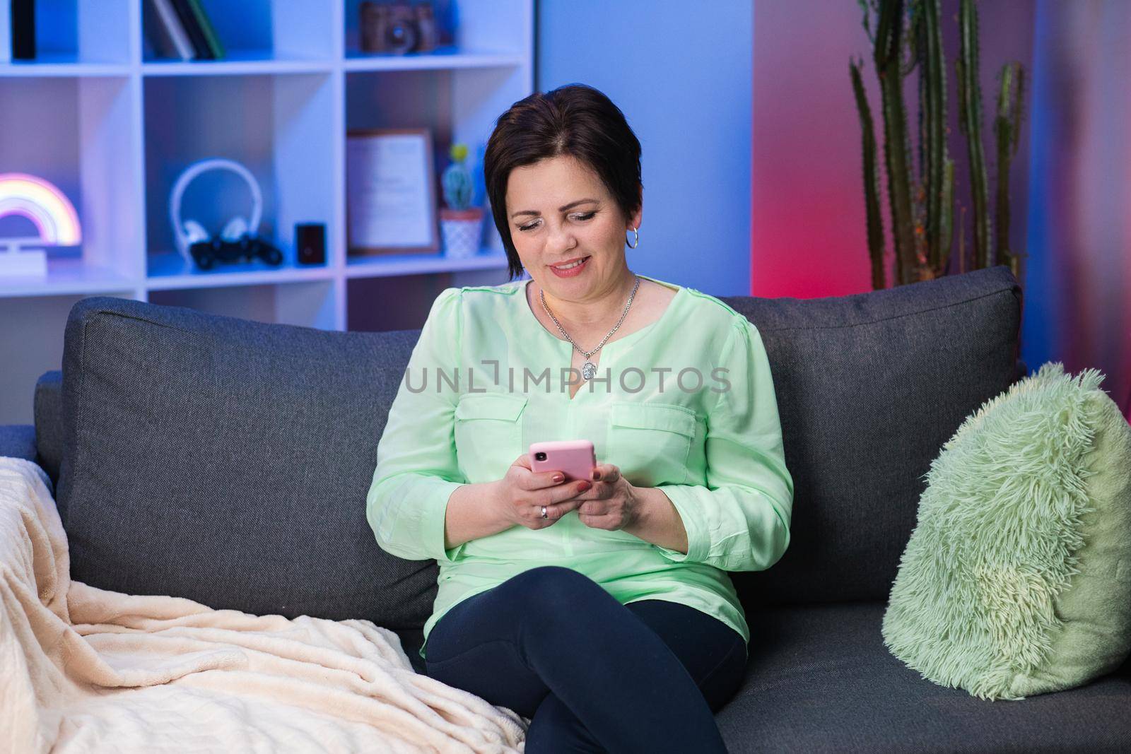 Senior Caucasian Woman with Short Black Hair Sitting on Couch at Home, Holding Invisible Smartphone in Hand and Chatting with Someone