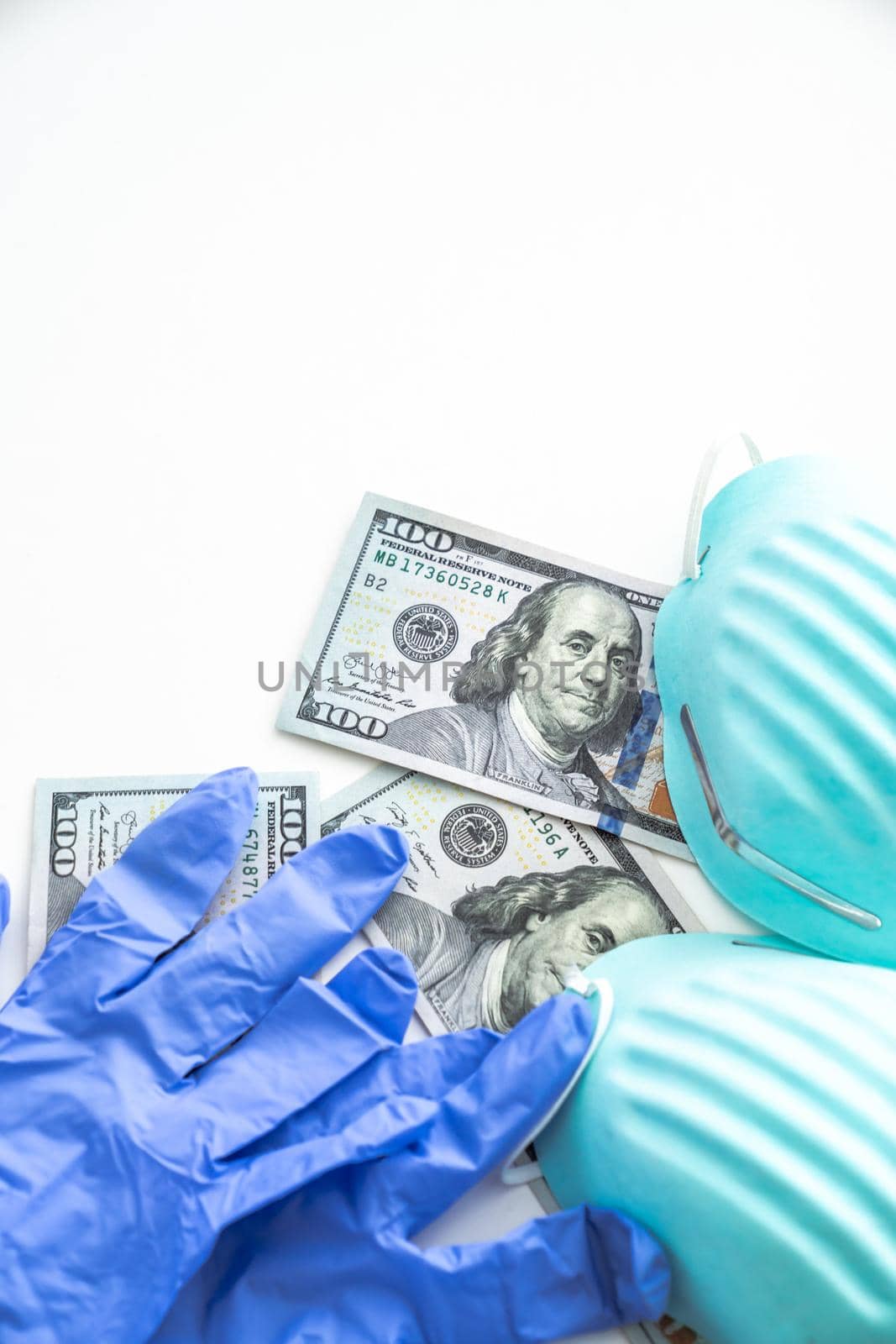 A close up of a few hundred dollar bills or cash laying below blue ribbed medical masks and pair of worn used latex gloves for protection from the coronavirus COVID-19 pandemic with white copy space.