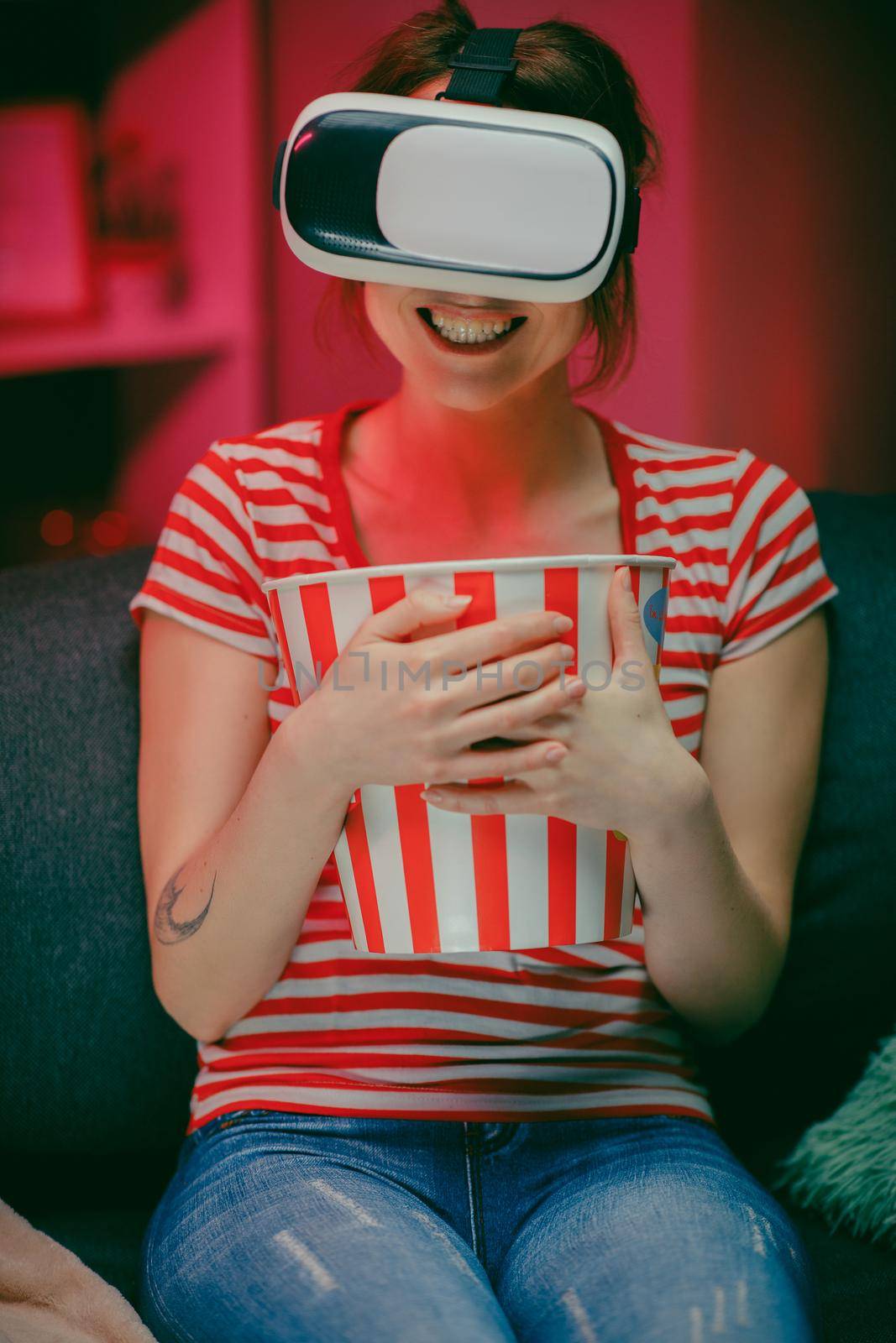 Woman wear VR headset and watch movie with popcorn at night. American woman sitting on the sofa in the VR glasses and watching something while eating popcorn. Indoor