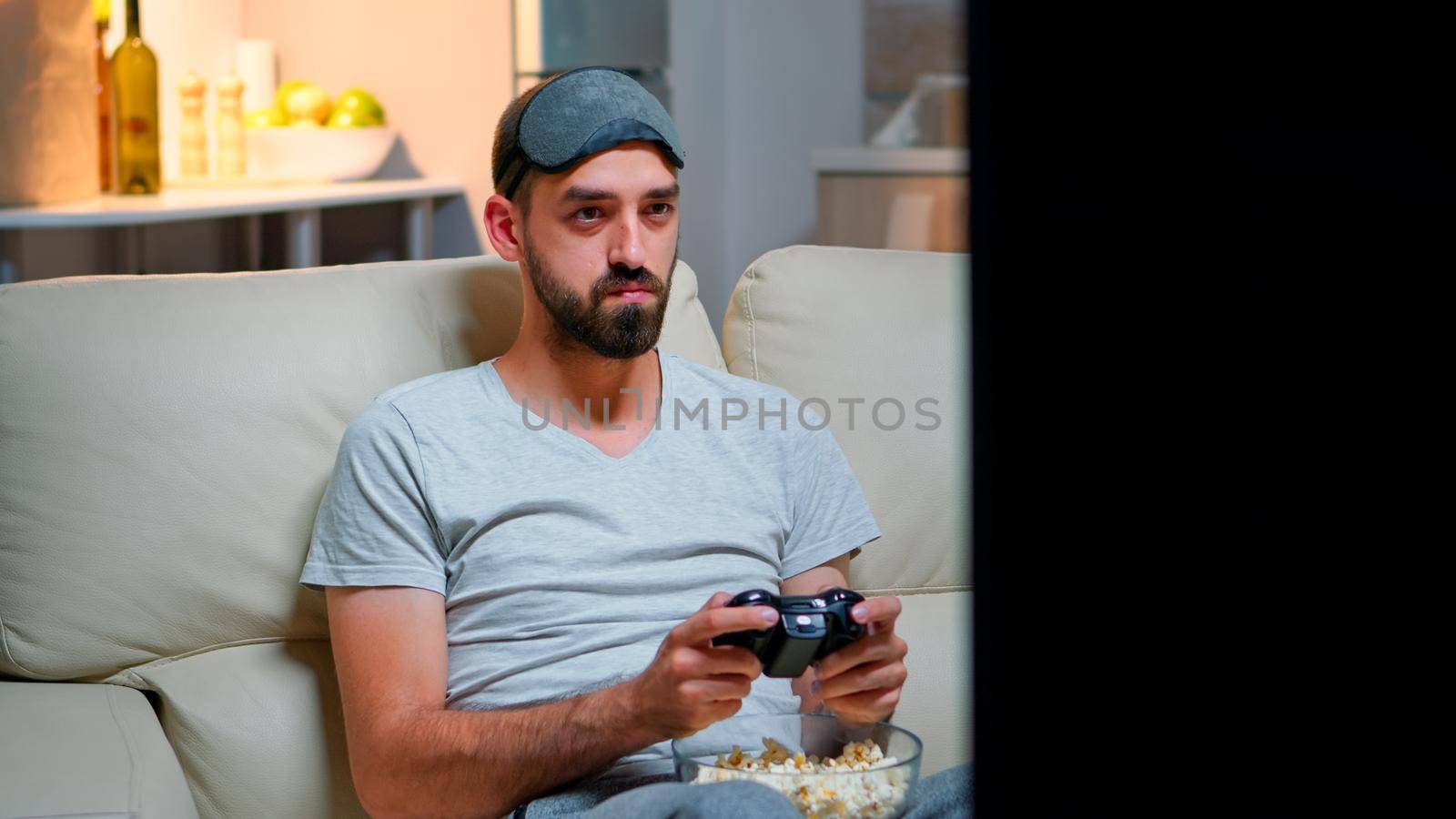 Close up of man with eye sleep mask playing videogames with joystick for gaming competition. Caucasian male sitting in front on television on sofa late at night in kitchen