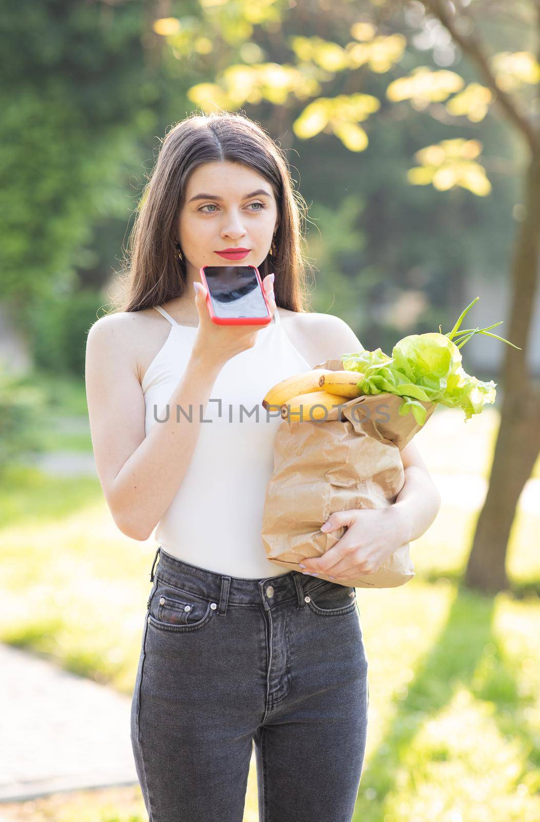 Happy girl using a smart phone voice recognition audio ai message speech function on line walking on a park background and carrying bag with food by uflypro