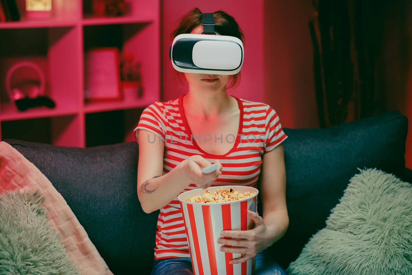 Portrait of the young woman sitting on the couch and having the VR headset, watching something while eating popcorn and smiling. Indoors
