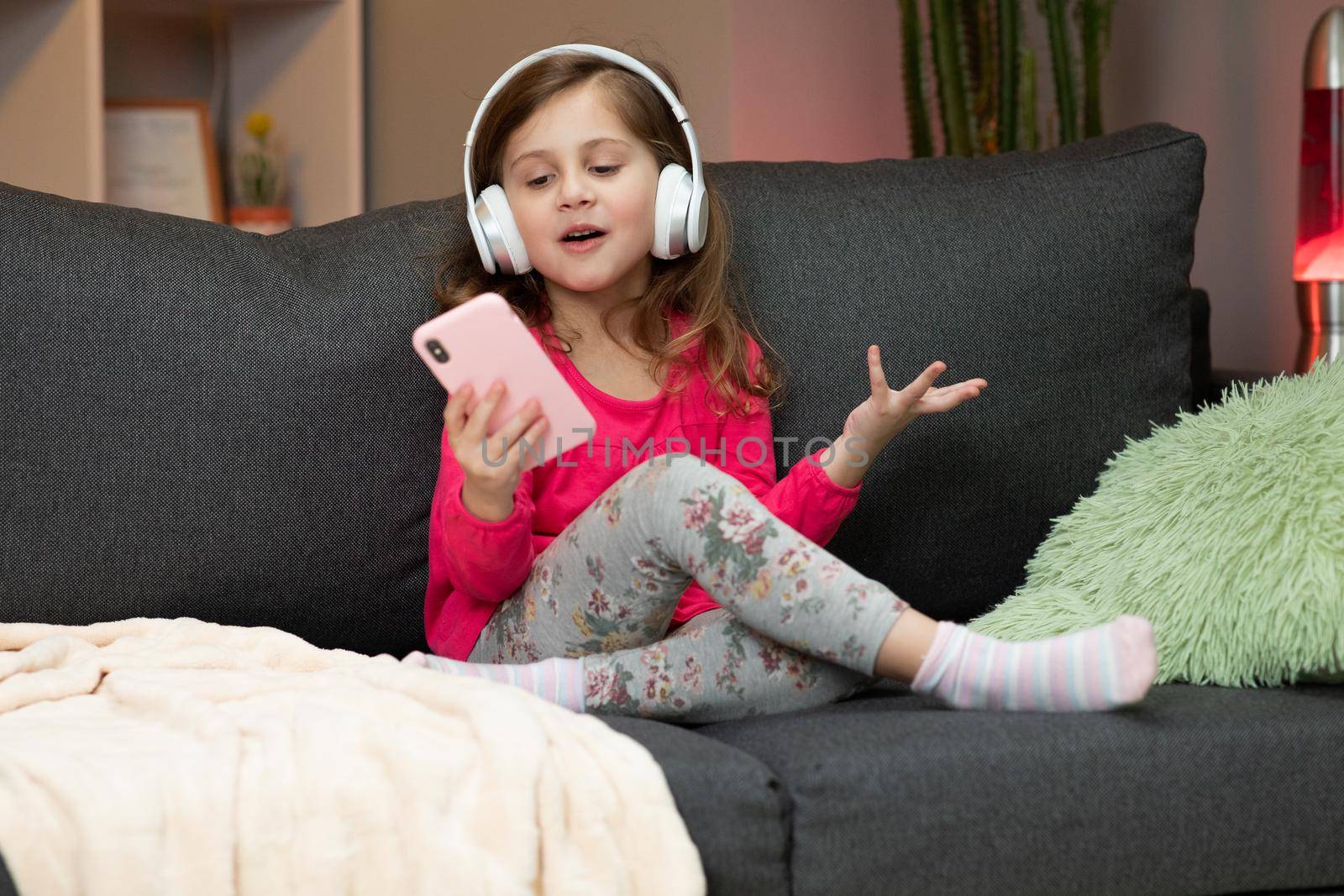 Little joyful girl in headphones is dance on sofa and using smartphone touching screen. Modern technology and happy childhood concept.