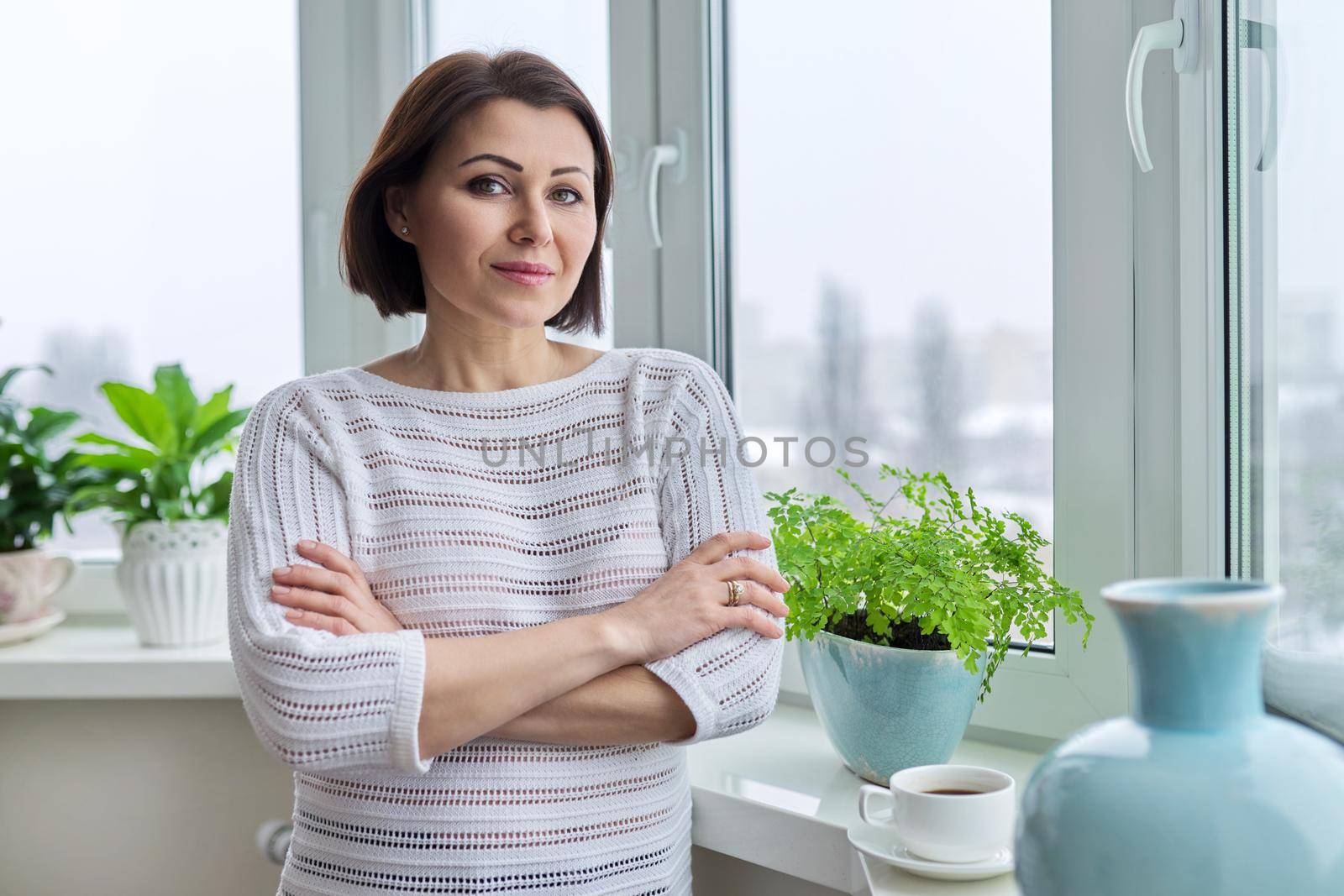 Portrait of smiling confident woman 45 years old. Middle aged positive woman with crossed arms looking at camera, standing at home near window, copy space. Mature people, health, beauty concept