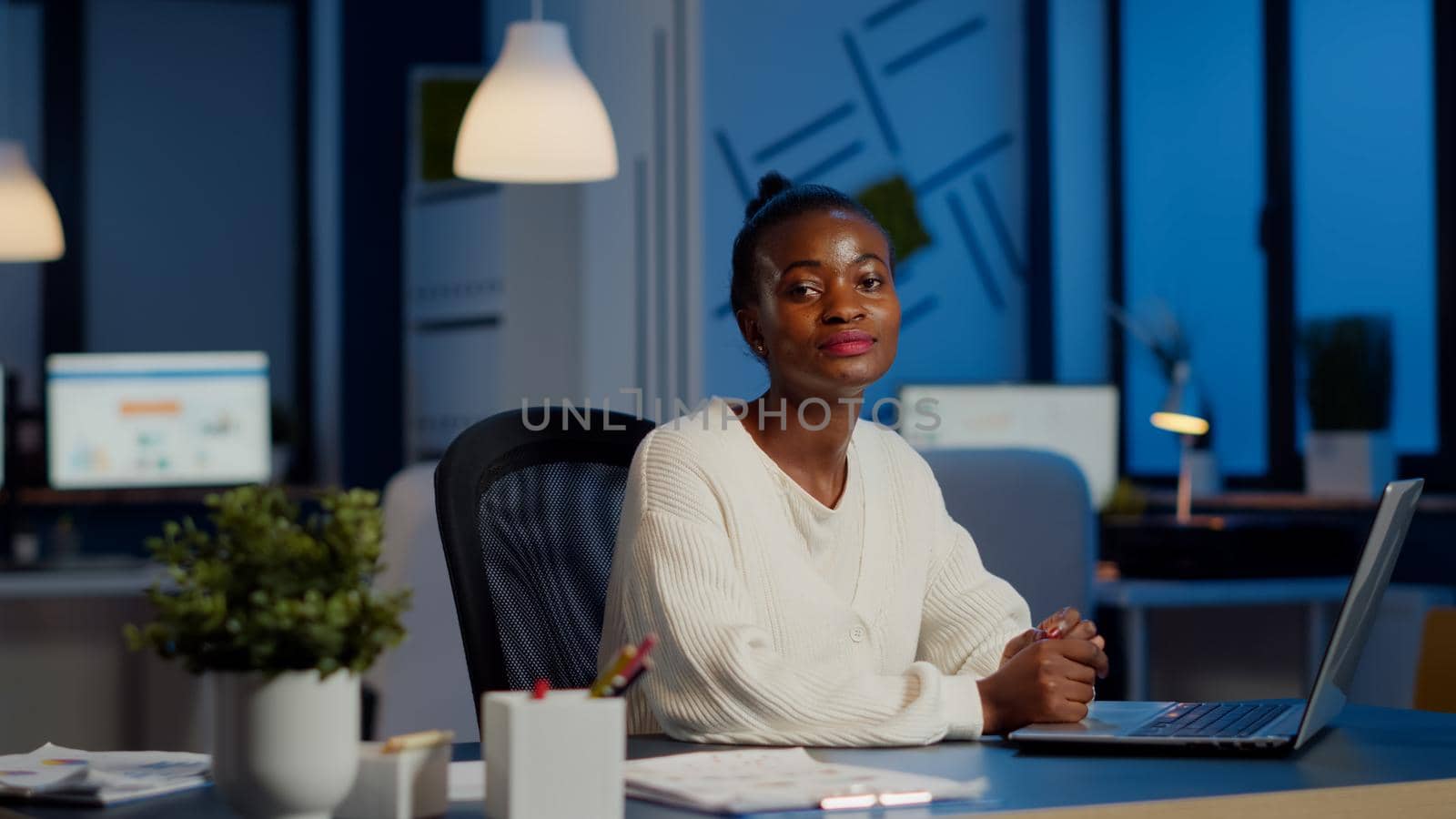 Tired black woman manager looking at camera sighing after typing on laptop, working late at night in start-up financial business office. Focused employee doing overtime respecting deadline of project
