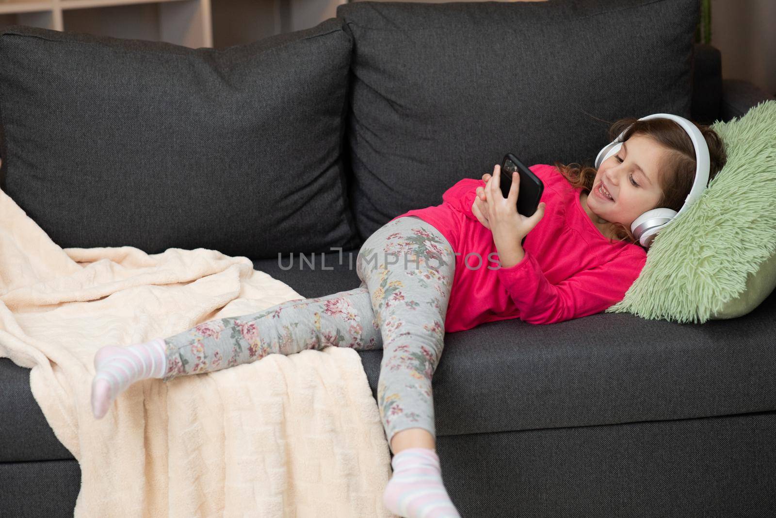 Happy Little girl in wireless headphones chilling laughing on sofa listening to favorite music holding phone using mobile online player app enjoy peaceful mood wearing earphones at home by uflypro