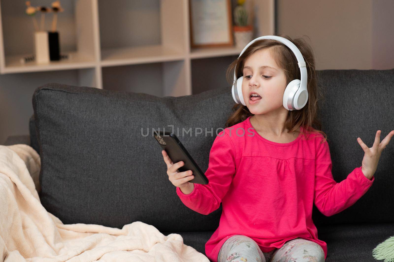 Little happy girl dancing on sofa while listening music in headphones at home. Girl wearing headphones dancing, singing and moving to the rythm by uflypro