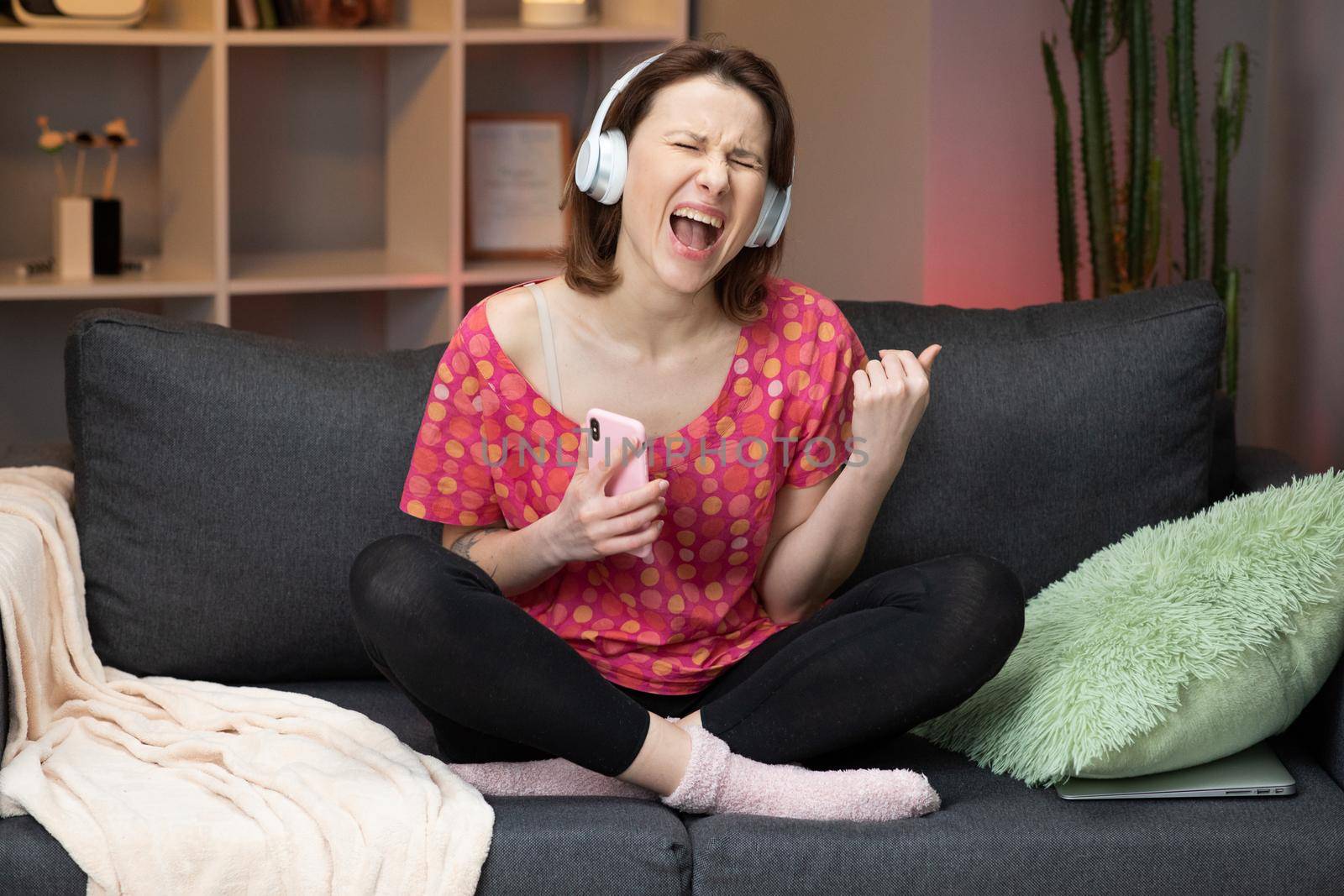 Caucasian girl sitting on sofa in home, smiling and listening to music in headphones on modern smartphone
